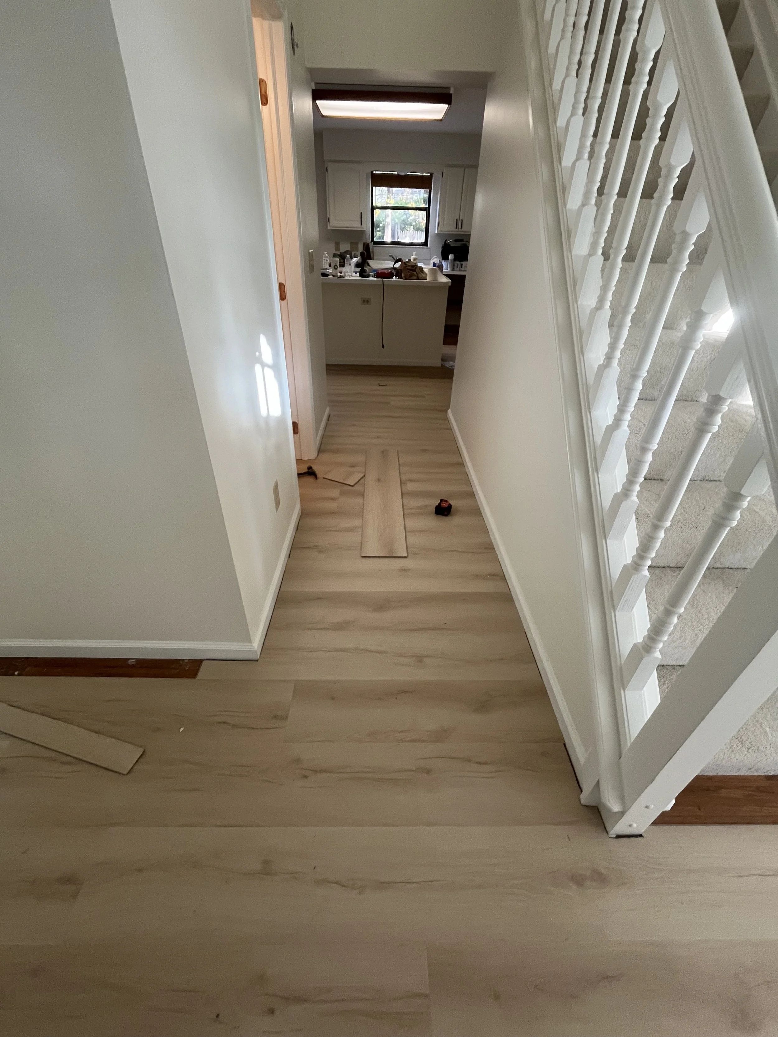 View of a hallway with light wood flooring, leading to a kitchen at the back with white cabinets and a window, and a staircase on the right side with white spindles. There are tools and materials on the floor and countertops, indicating ongoing home 