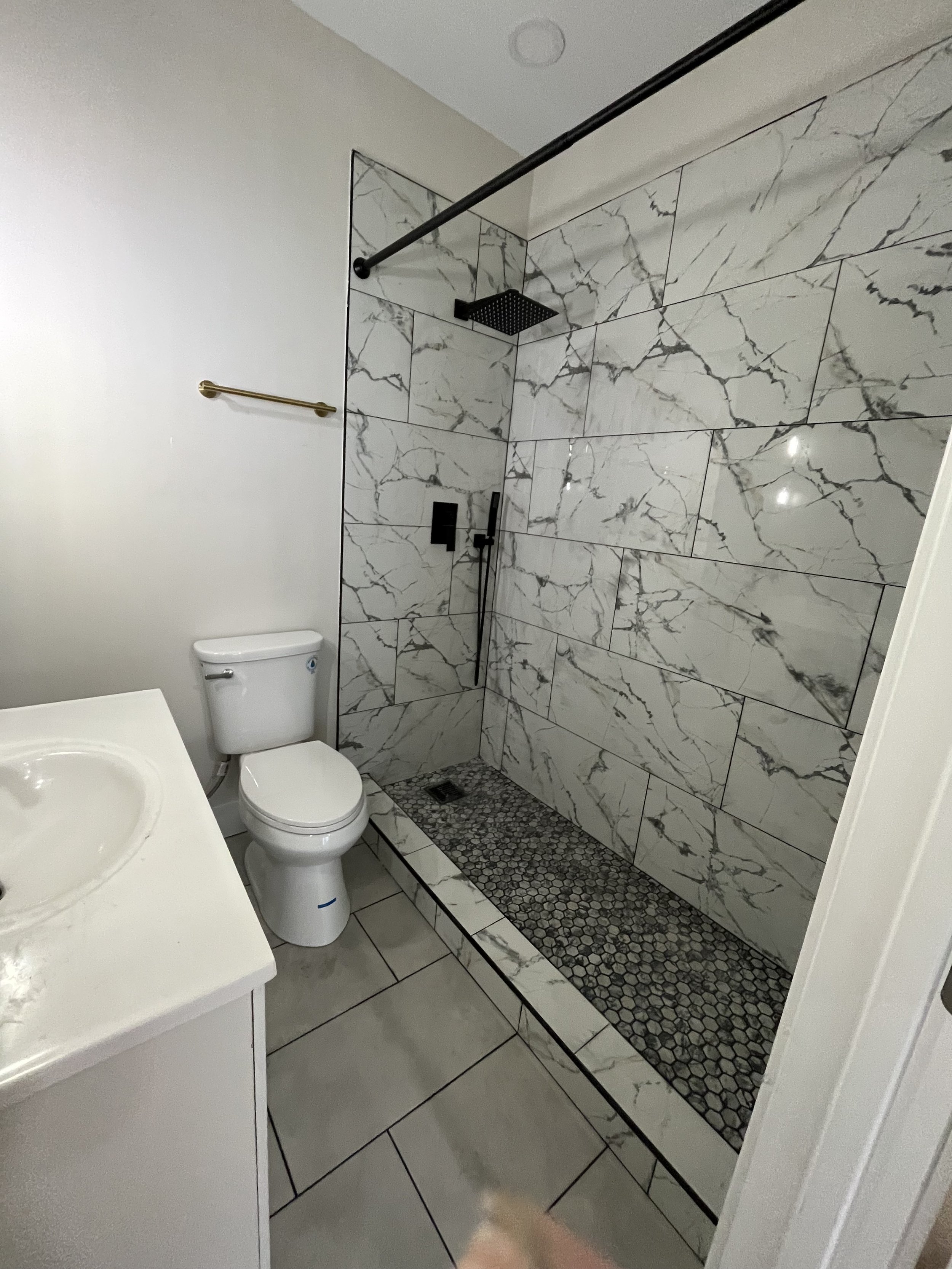 Bathroom with a walk-in shower featuring marble tiles and pebble flooring, a white toilet, a white vanity with a sink, a gold towel bar on the wall, and a black shower fixture with a rainfall and handheld showerhead.