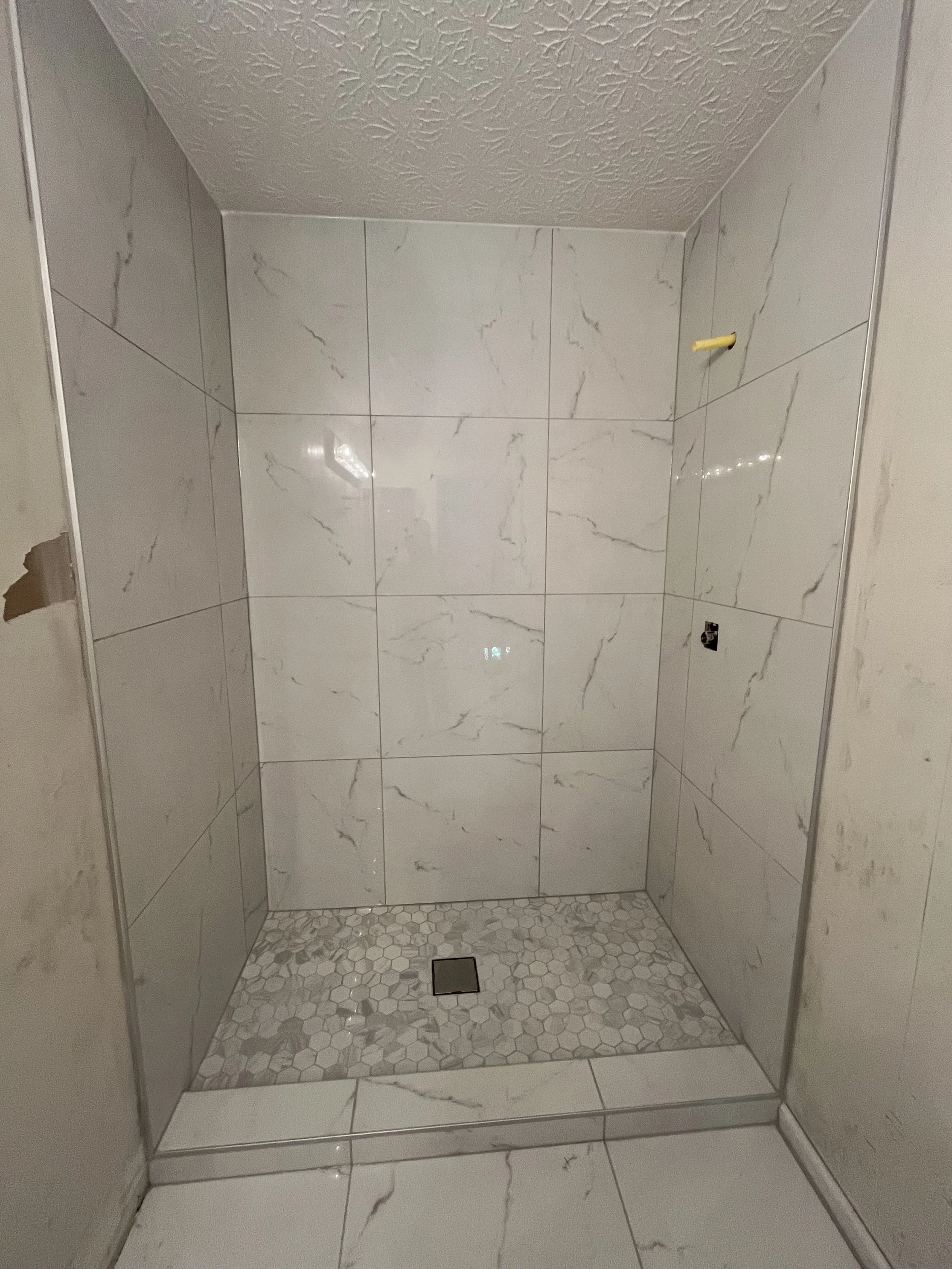 Empty shower stall with white marble tiles on the walls and small hexagonal tiles on the floor, with a drain in the center.