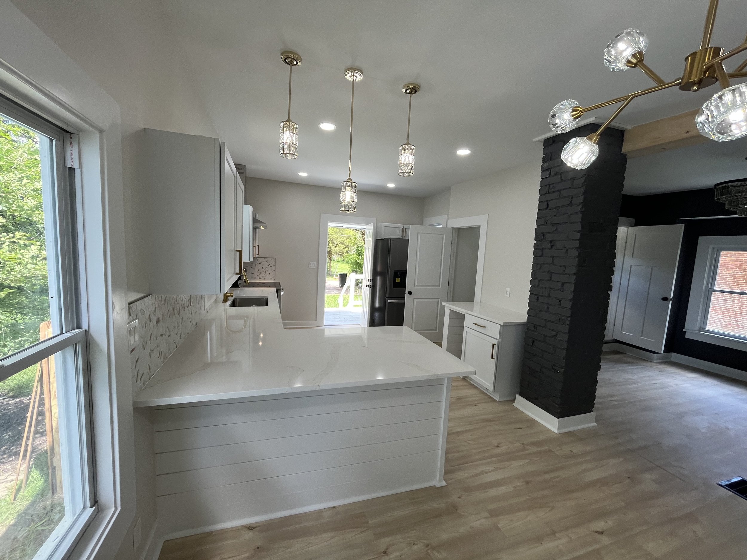 Modern kitchen with white cabinets, marble countertop island, black brick column, pendant lights, large windows, and hardwood flooring.