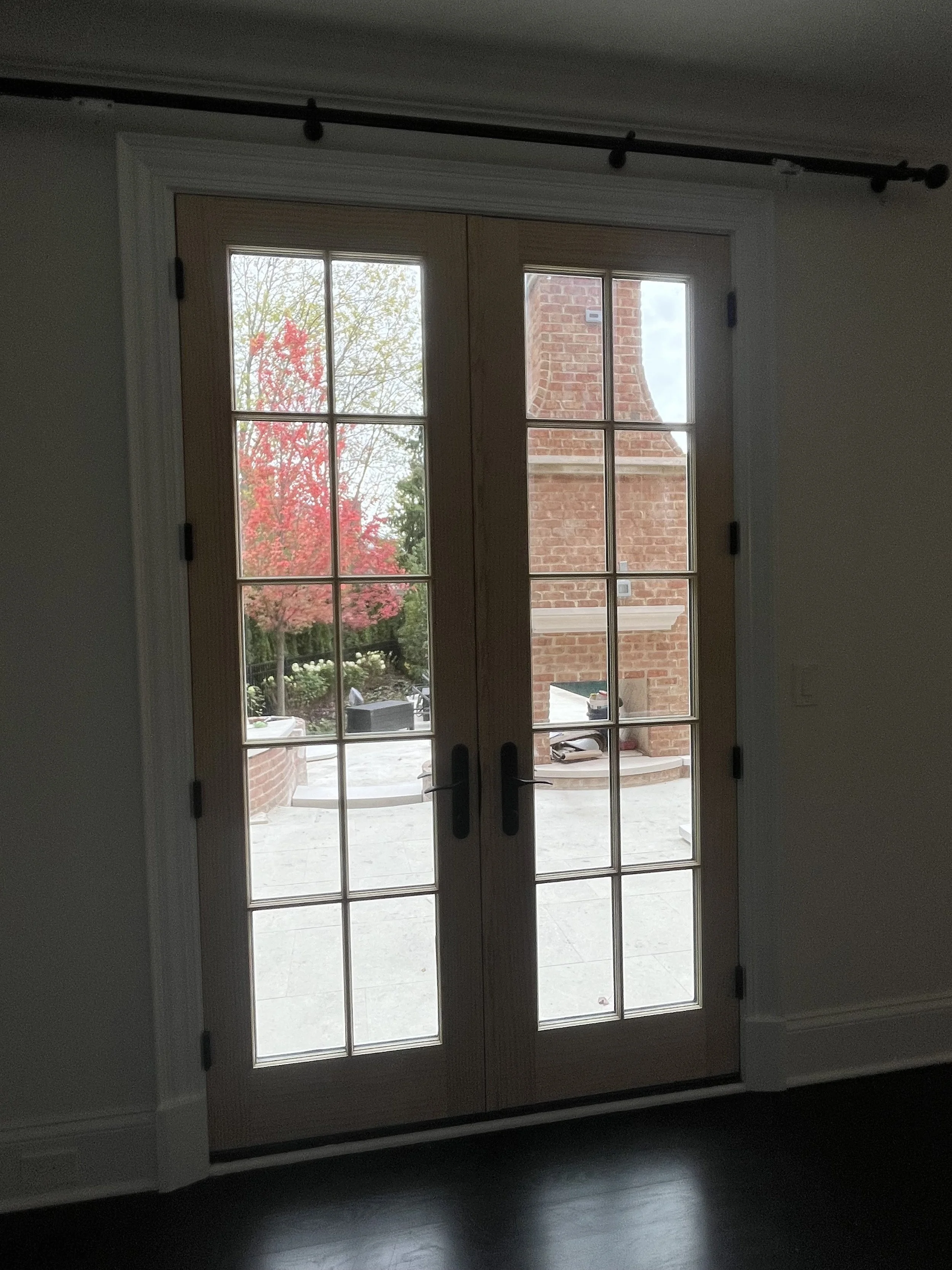 View through glass-paneled double doors showing a backyard with a tree with red leaves, a brick chimney, and potted plants.