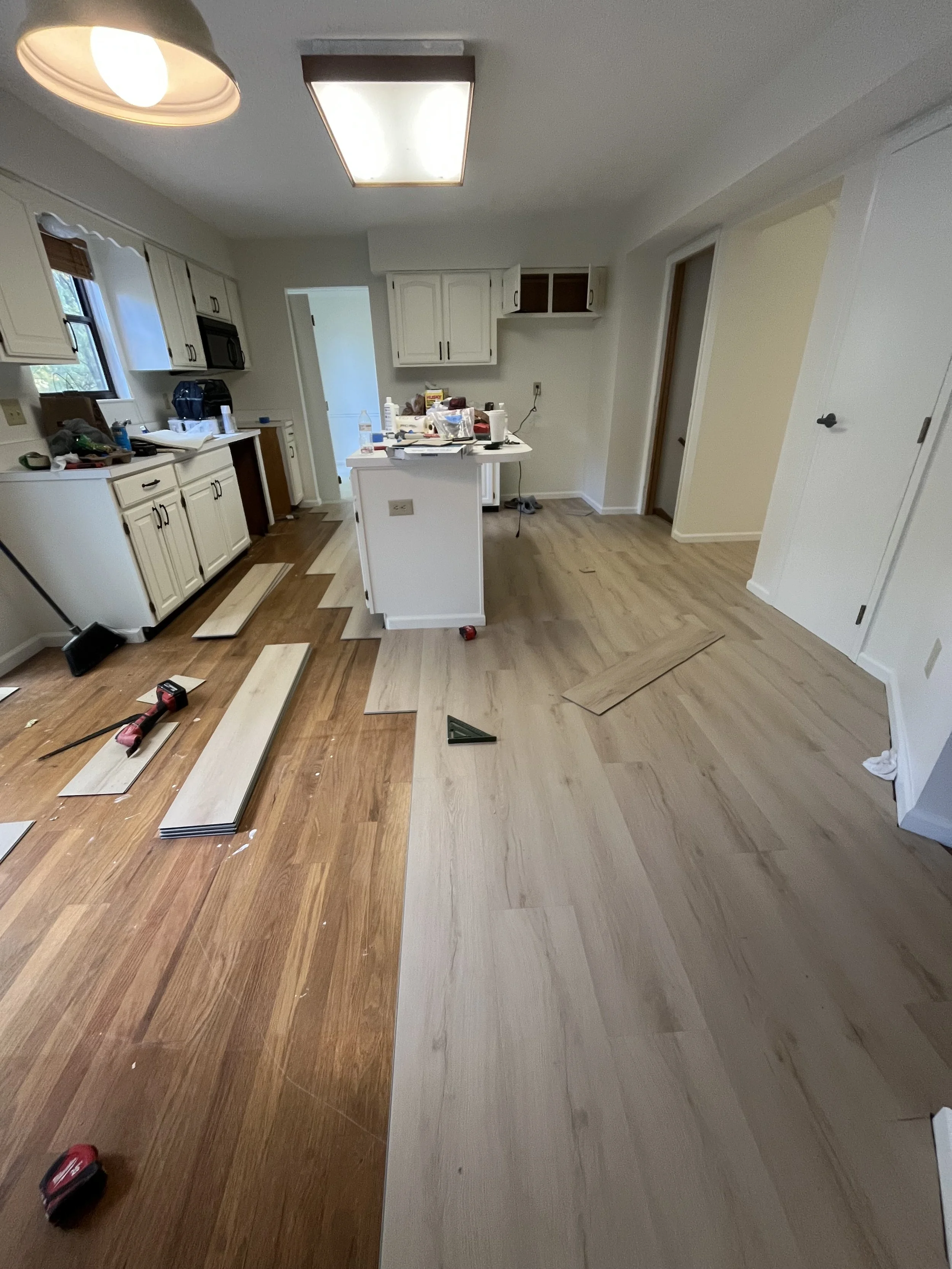 Kitchen undergoing flooring renovation, with partially installed new hardwood flooring, tools, and materials scattered around.