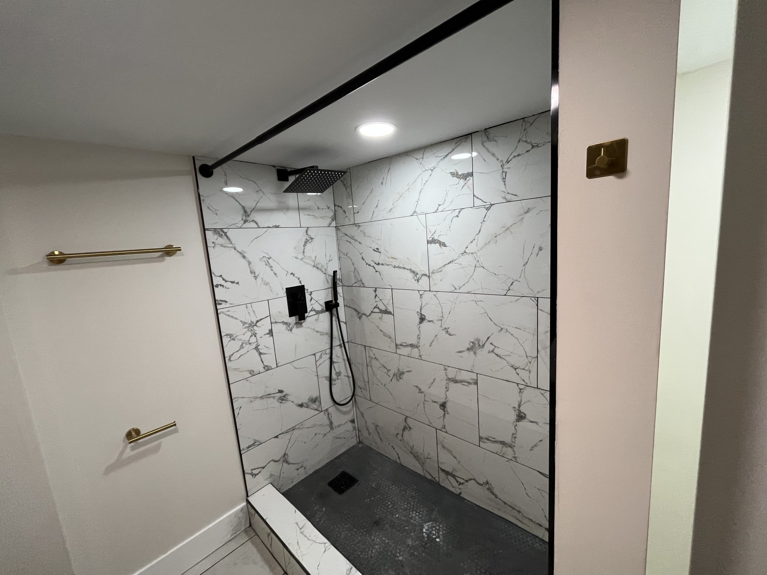 Modern walk-in shower with white marble tiles, a black rain showerhead, black handheld shower, and black controls, with gold towel bars on adjacent wall.