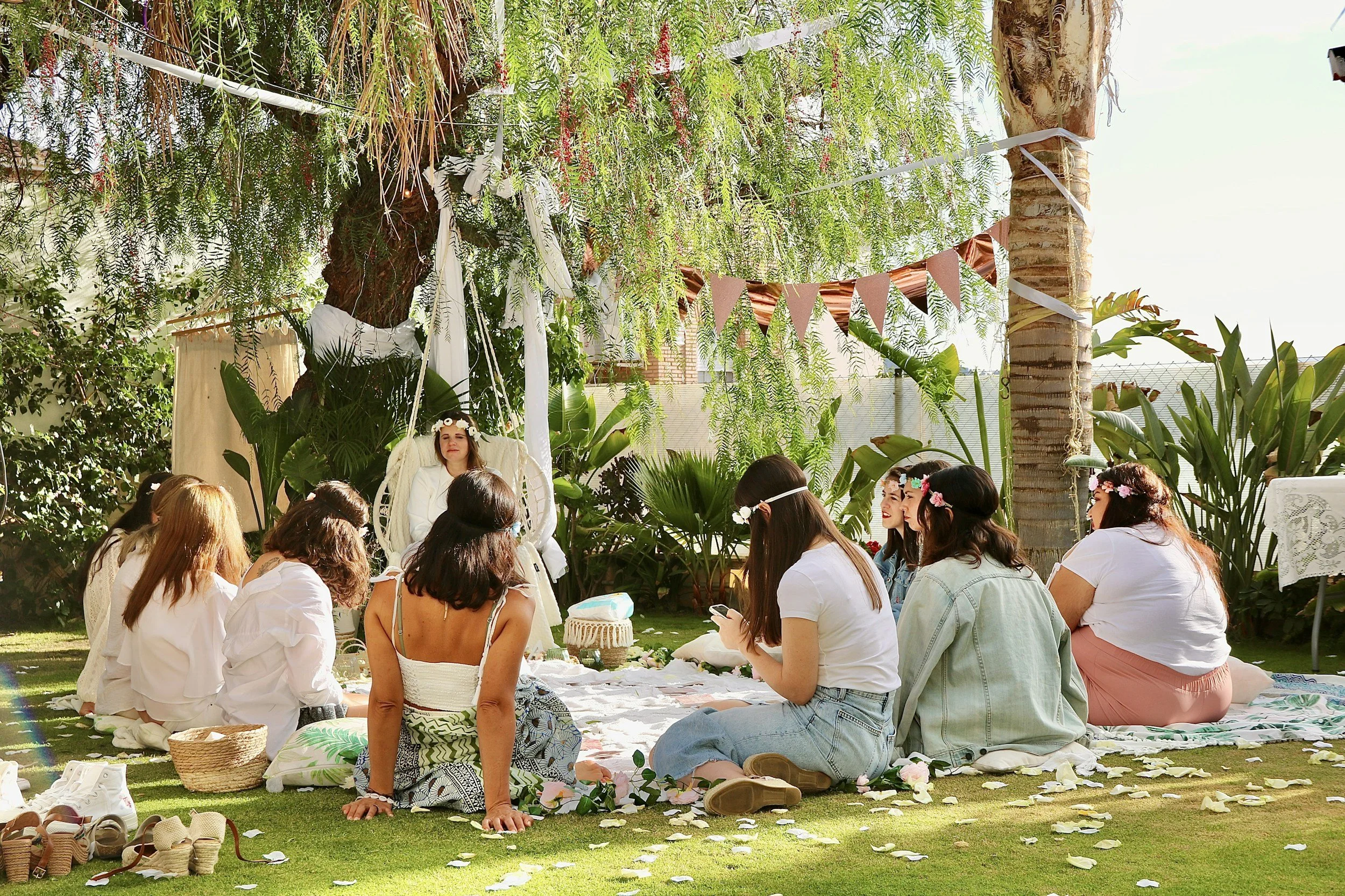 People sitting on a blanket in a lush garden, participating in a celebration or gathering with a woman dressed as an angel, surrounded by trees and hanging bunting flags.