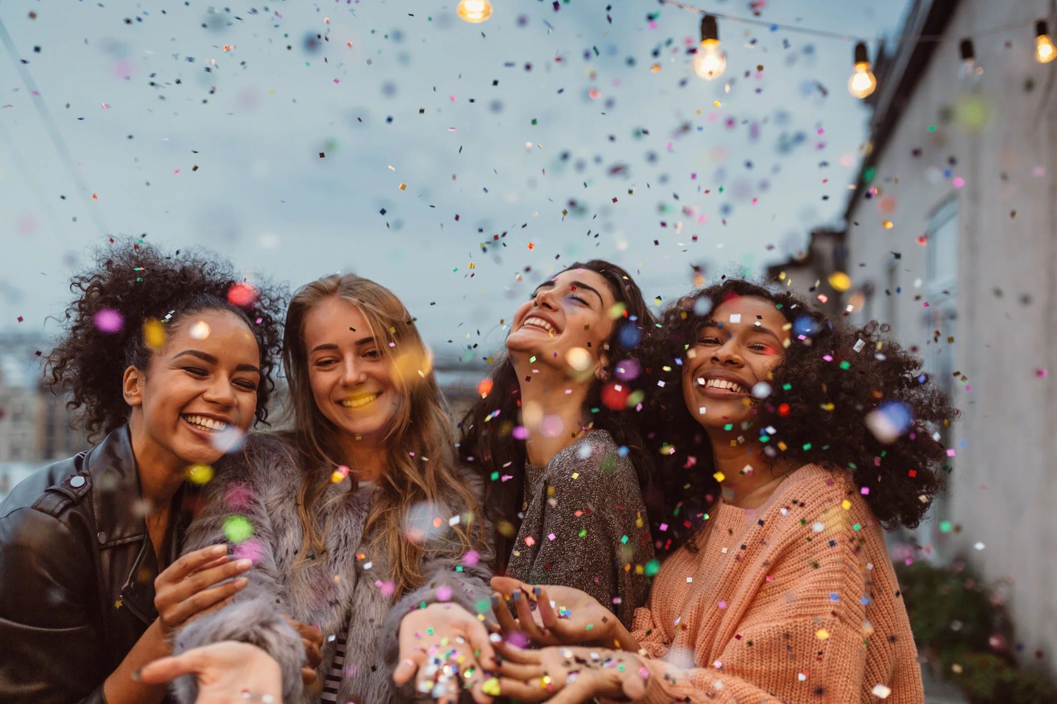 Four women celebrating outdoors with colorful confetti falling around them, smiling and enjoying the moment, during daytime with string lights overhead.