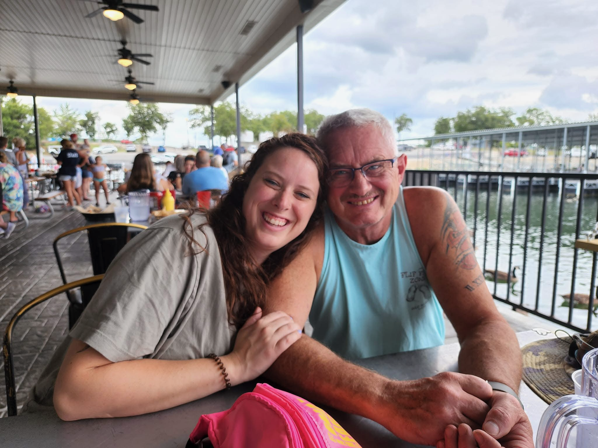 Two people smiling happily, a woman with dark curly hair and a man with short gray hair and glasses, sitting at an outdoor restaurant or cafe with a waterfront view, other people in the background, and cloudy skies.