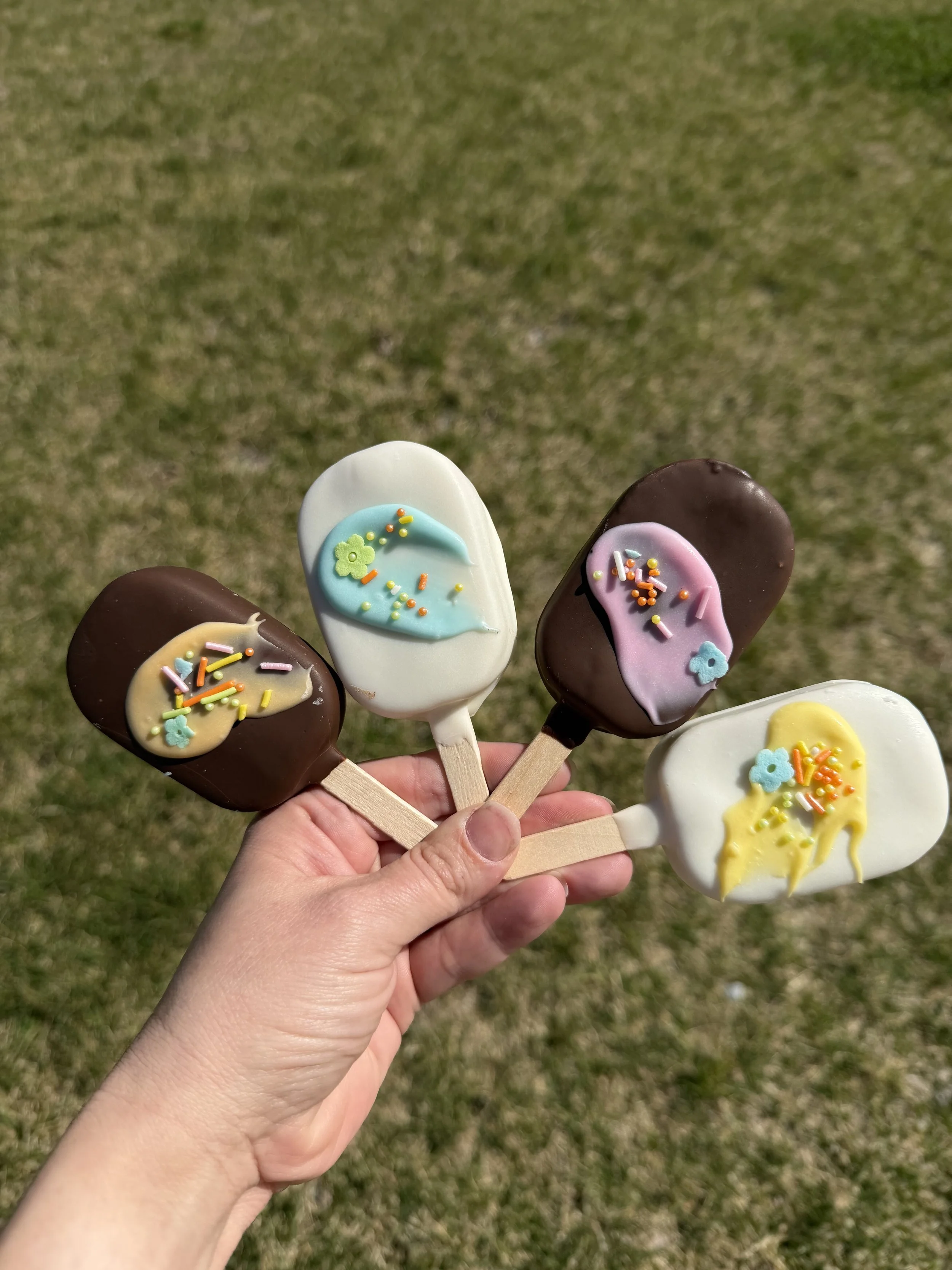 Person holding five colorful ice cream-shaped popsicles with sprinkles against a grassy background.