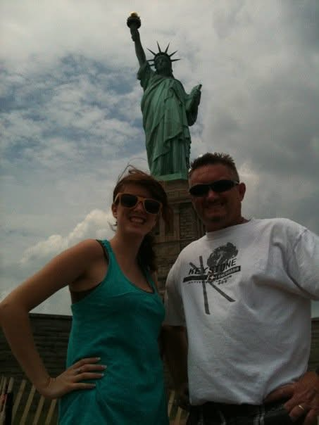 A woman and a man posing in front of the Statue of Liberty on a cloudy day, with the statue holding a torch and wearing a crown.