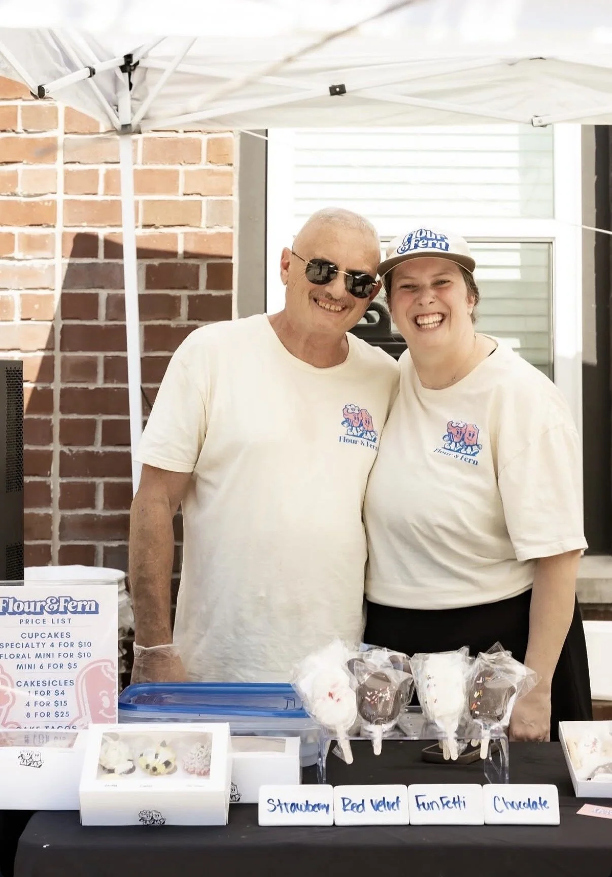 Two smiling people wearing ivory t-shirts with a pink and blue logo for Flour & Fern, standing behind a table with decorated cake pops labeled strawberry, red velvet, funfetti, and chocolate, under a white canopy with brick wall in the background.