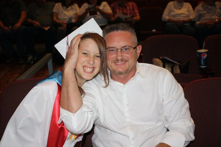 A young woman in a graduation cap and gown sitting next to a man in a white shirt, smiling at a graduation ceremony.