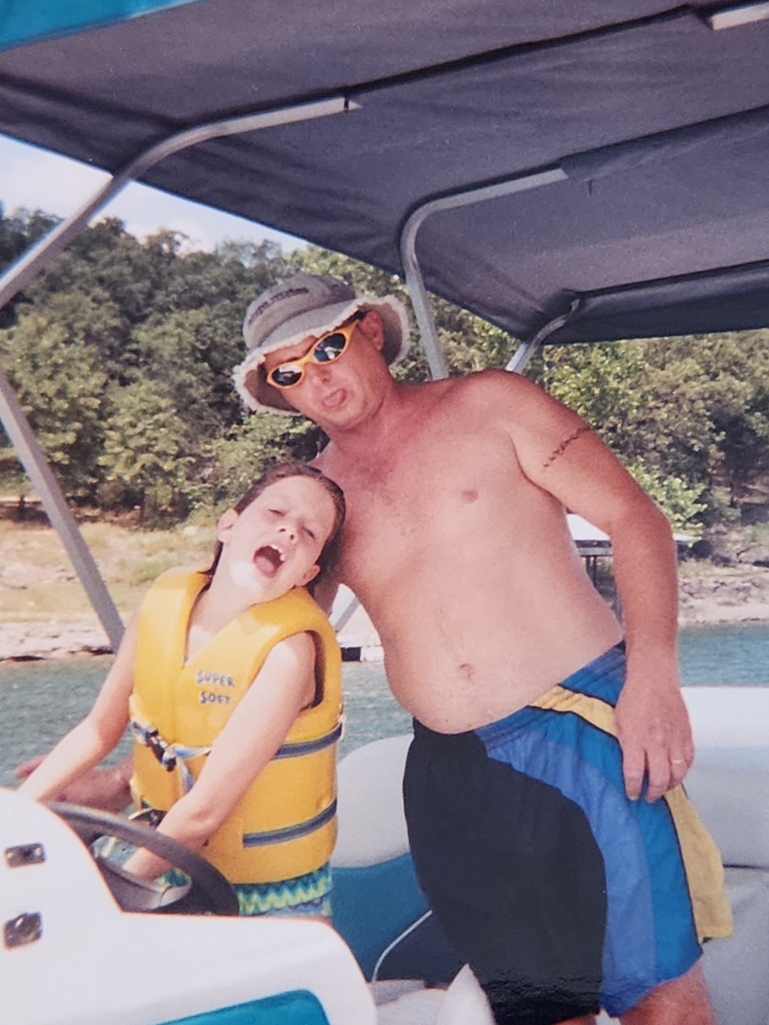 A shirtless man wearing a sun hat, sunglasses, and swim trunks standing next to a young boy in a yellow life jacket at the beach under a beach umbrella.