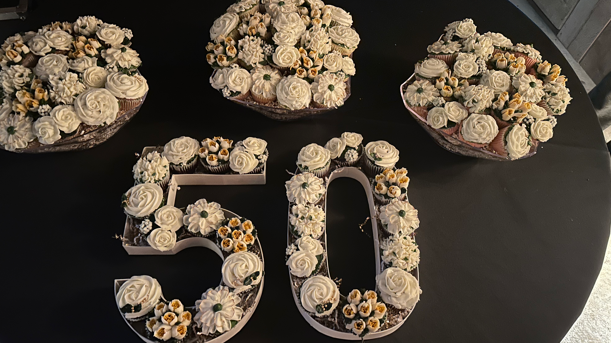 Multiple cakes decorated with white and yellow flowers on a dark table.