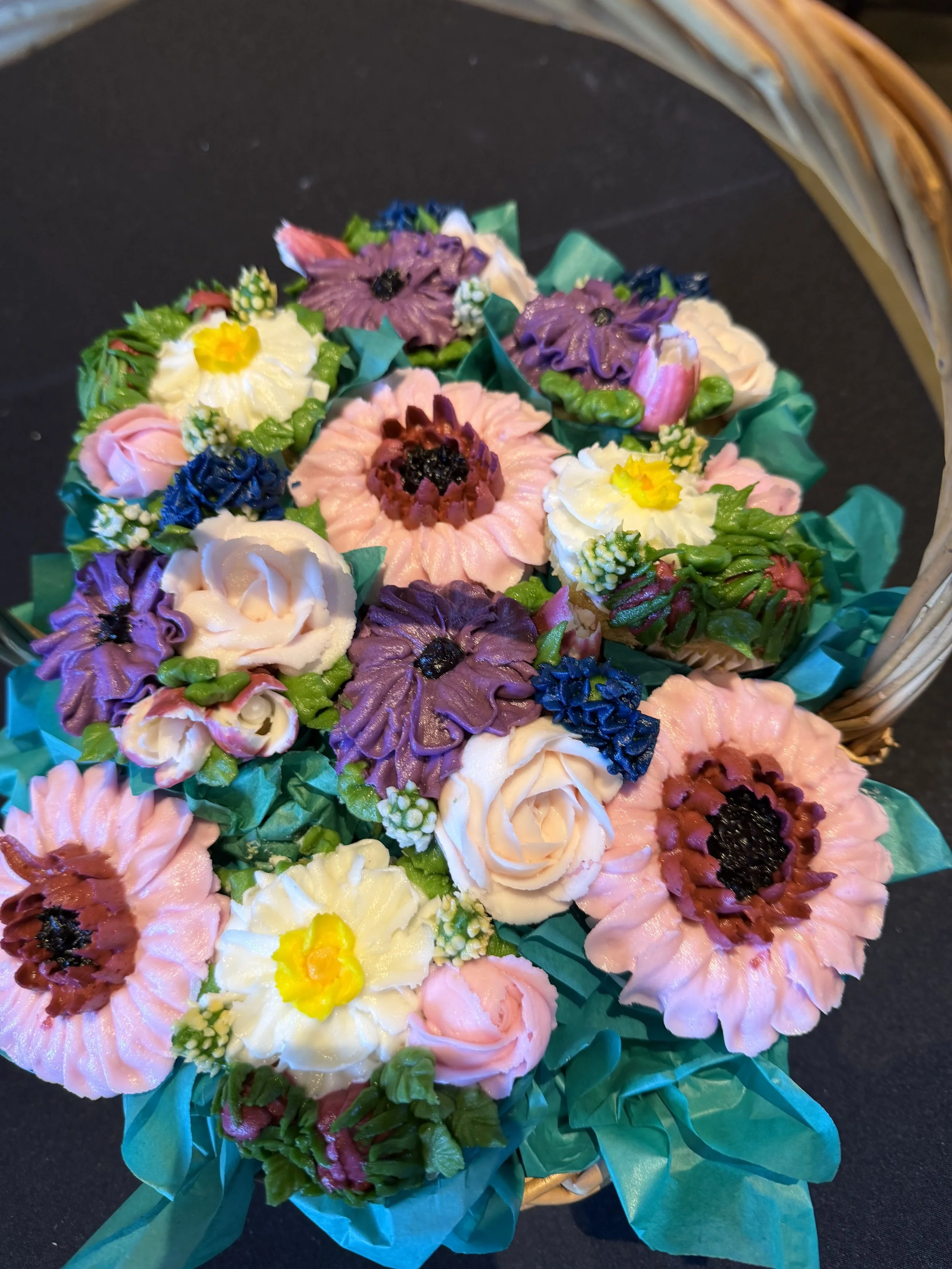 A basket of colorful handcrafted flower-shaped cookies in various shades of pink, purple, white, yellow, and blue, with green paper tissue accents.