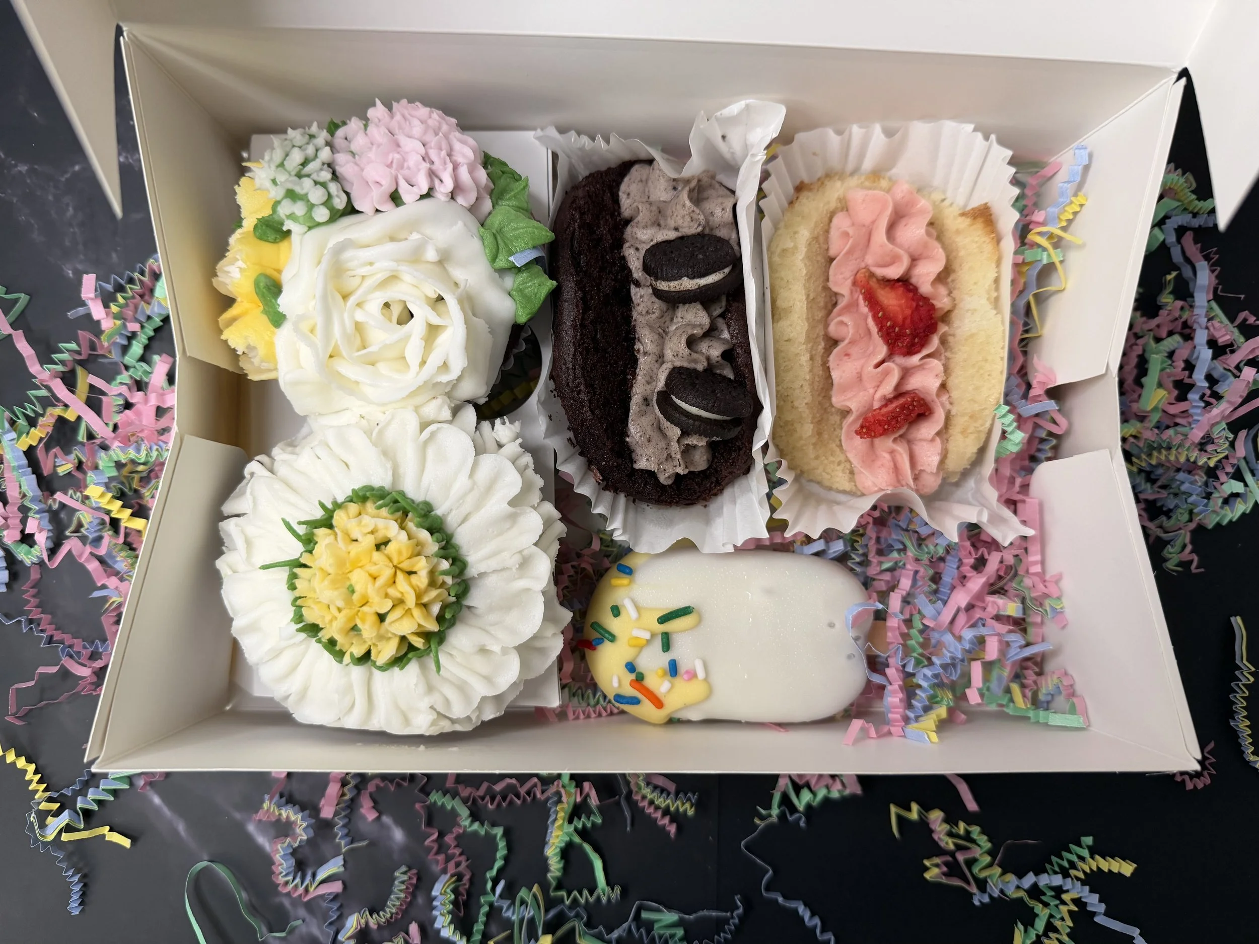 A white box with assorted decorated cupcakes, including flowers, cookies, and sprinkles, surrounded by colorful confetti and shredded paper.