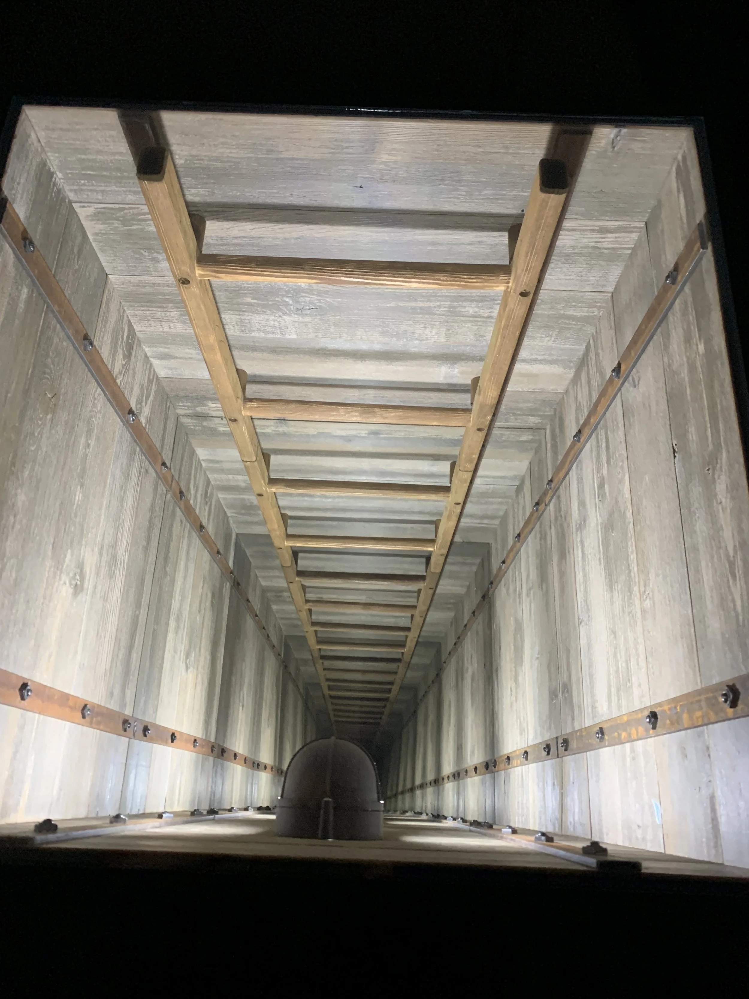 Looking down a deep well or shaft with a ladder attached inside, constructed with wood and metal.