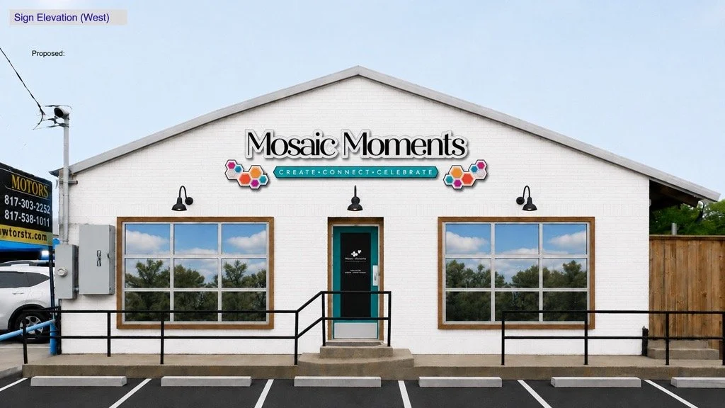 Front view of a white brick building labeled "Mosaic Moments" with windows reflecting trees, black outdoor lights, and a sign that says "Create, Connect, Celebrate."