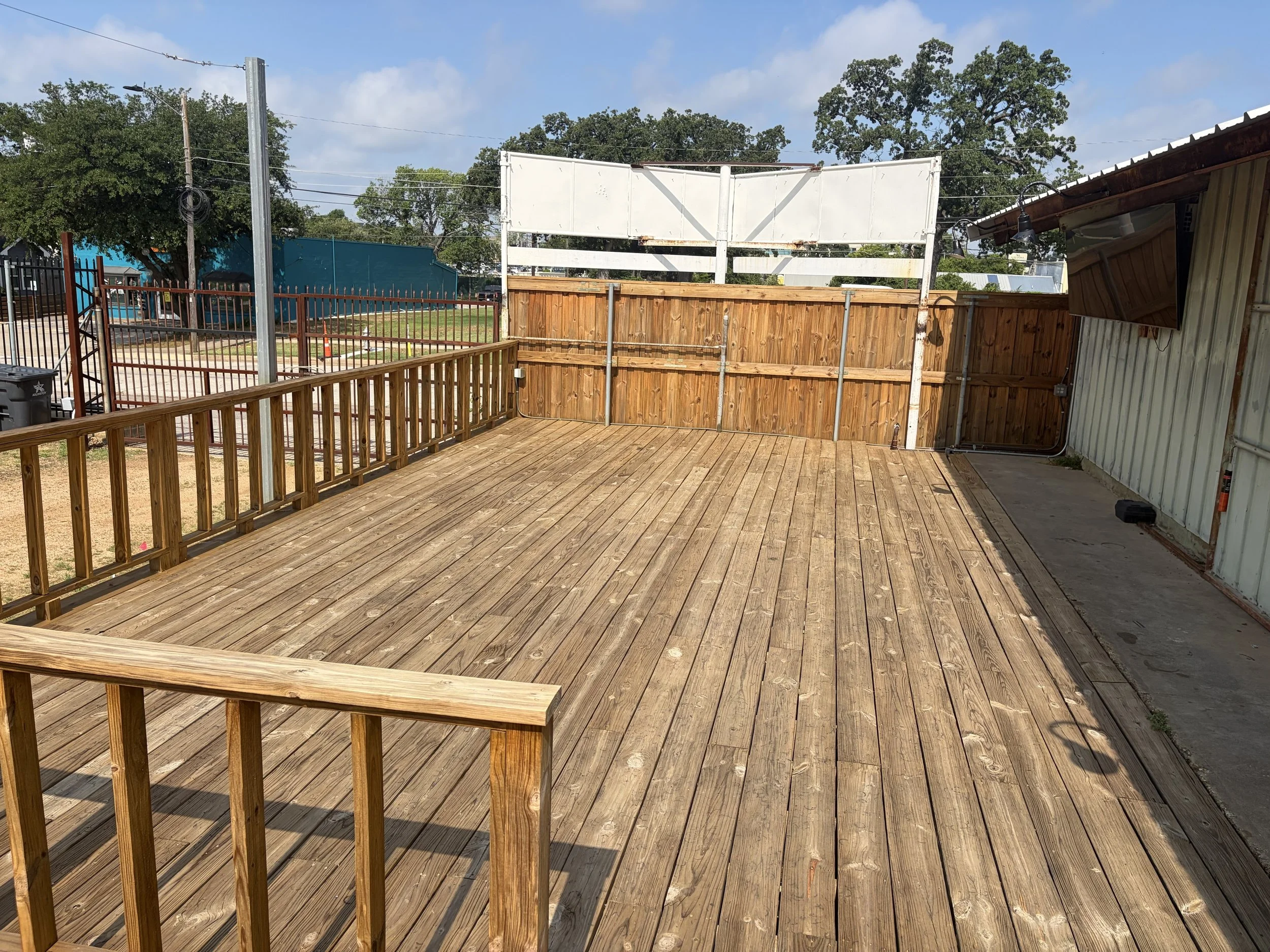 A newly constructed wooden deck with a safety railing, adjacent to a building with a metal roof and a wall-mounted TV. In the background, there is a backyard with trees, a chain-link fence, and a blue building. The sky is partly cloudy with blue skies and some clouds visible.