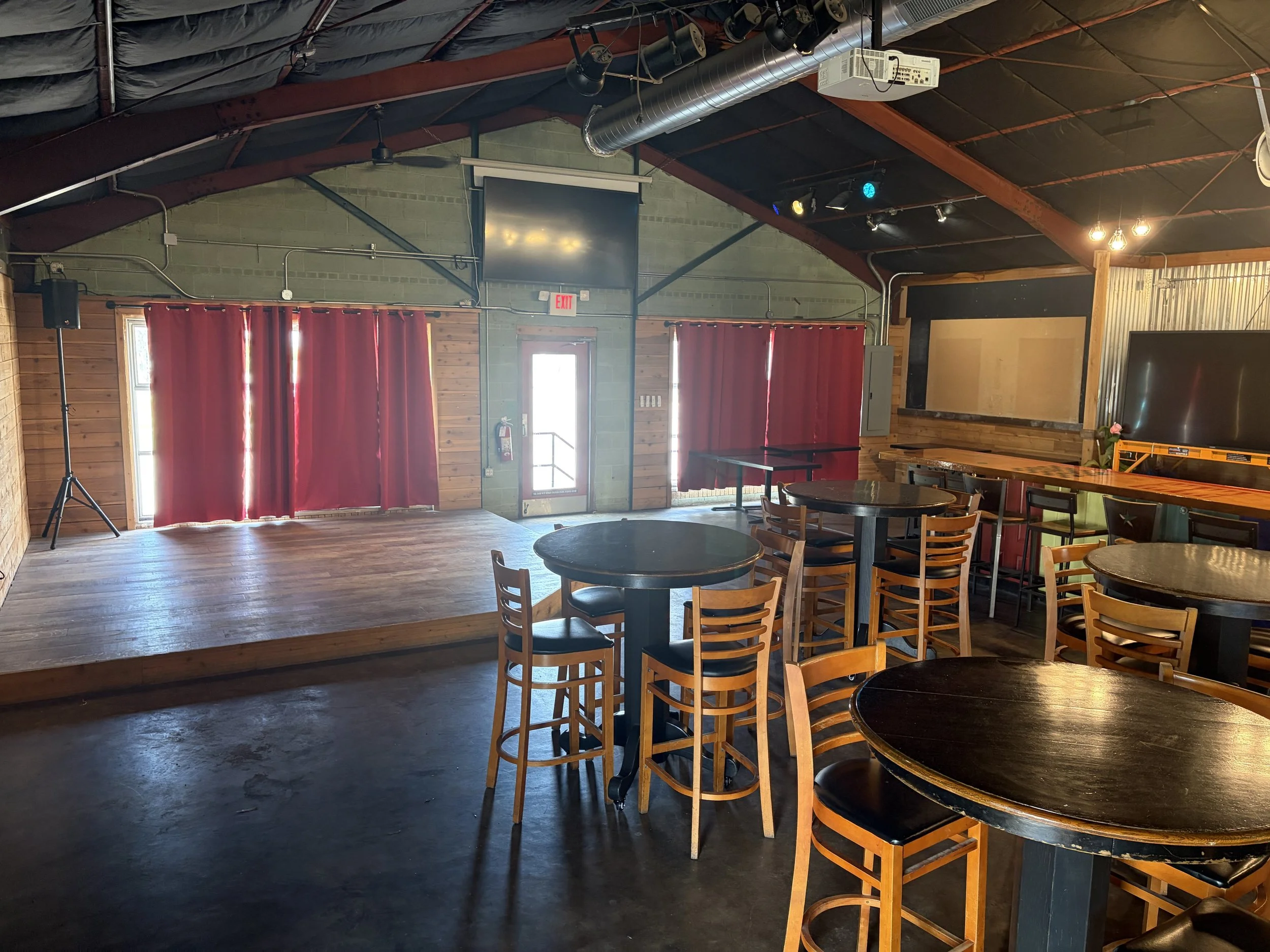 An empty event space or restaurant with wooden and metal interior design. Red curtains cover windows, and there are round tables with black chairs. Overhead lighting is warm, and there is a large monitor mounted on the wall. The ceiling has exposed ductwork and stage lighting.