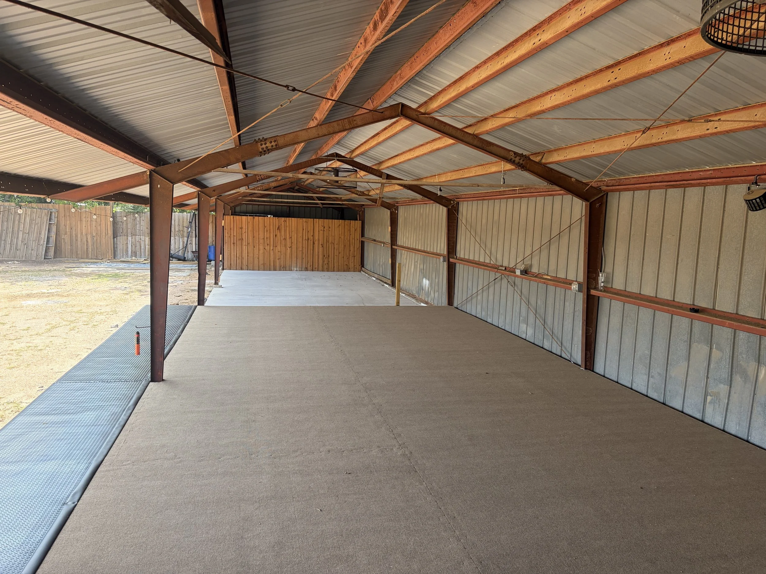 The image shows the interior of an empty metal garage or workshop with a concrete floor, wooden beams supporting a corrugated metal roof, and a wooden privacy wall at the back. There is also outdoor sunlight coming in from the open sides.