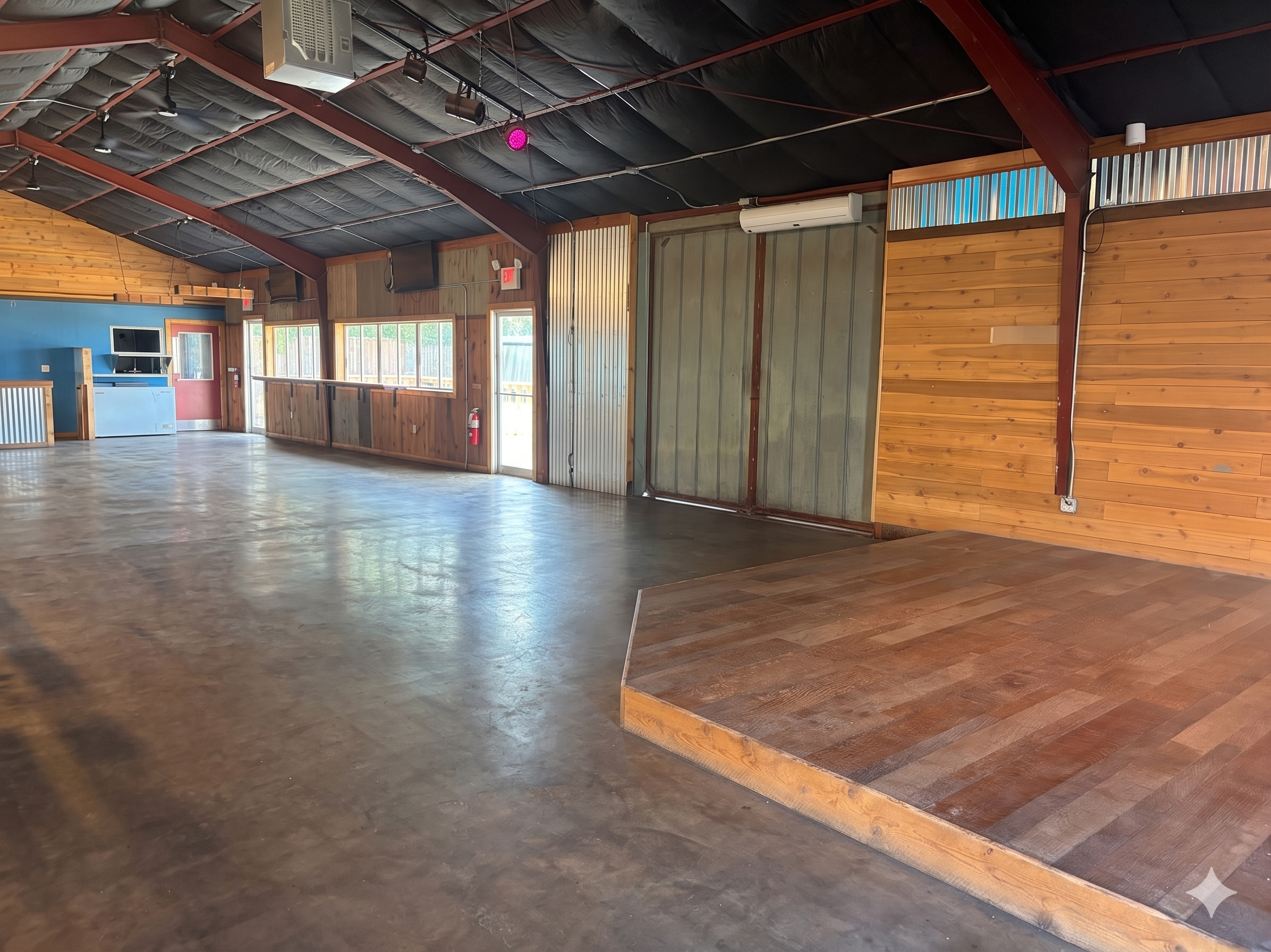 Interior of a wooden hall or dance studio with a polished floor, wooden paneled walls, and a black ceiling with stage lighting. There is a door with a glass window and an emergency exit sign, a television, and a suspended air conditioning unit.