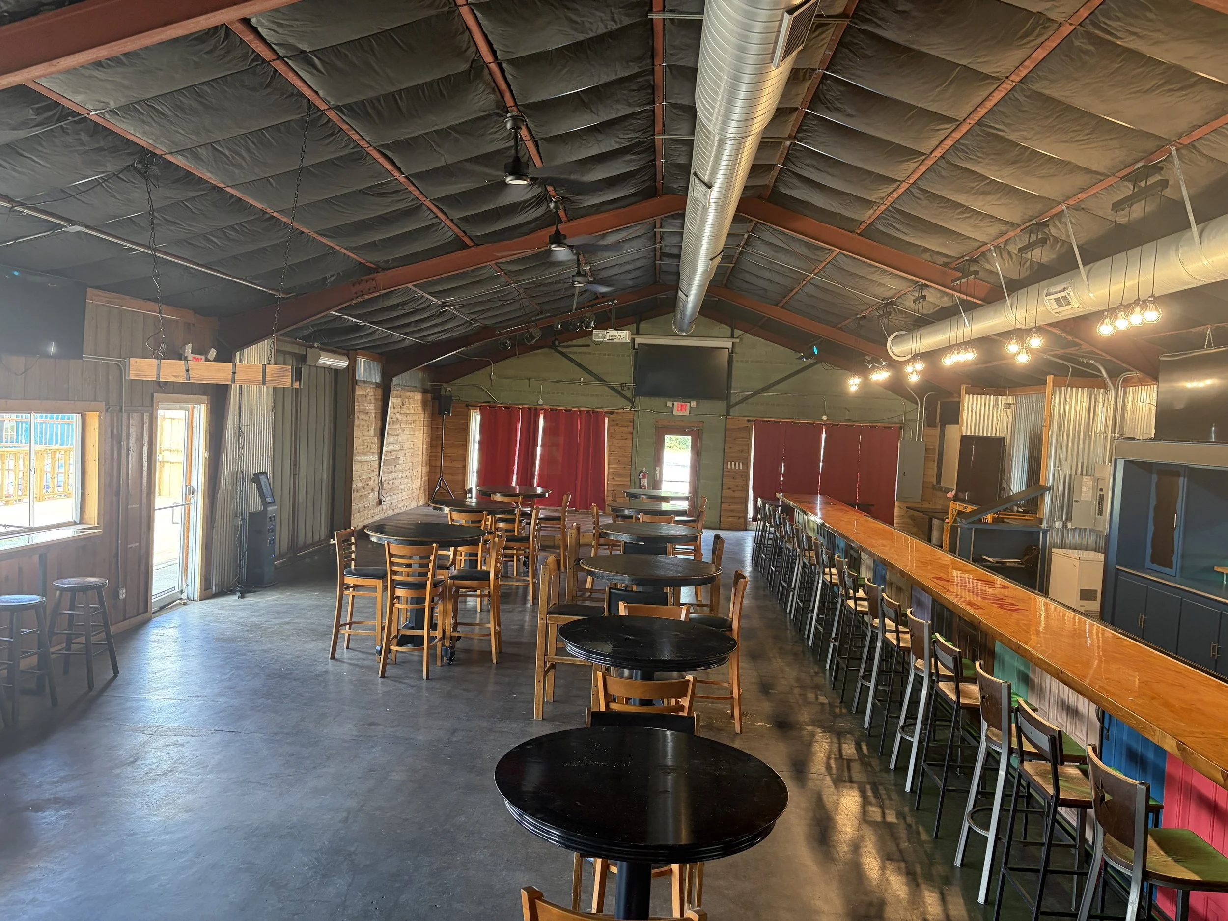 Interior of a restaurant or bar featuring wooden walls, a wooden bar counter with barstools, round black tables with chairs, a large ventilation duct on the ceiling, warm lighting, and red curtains covering the windows.