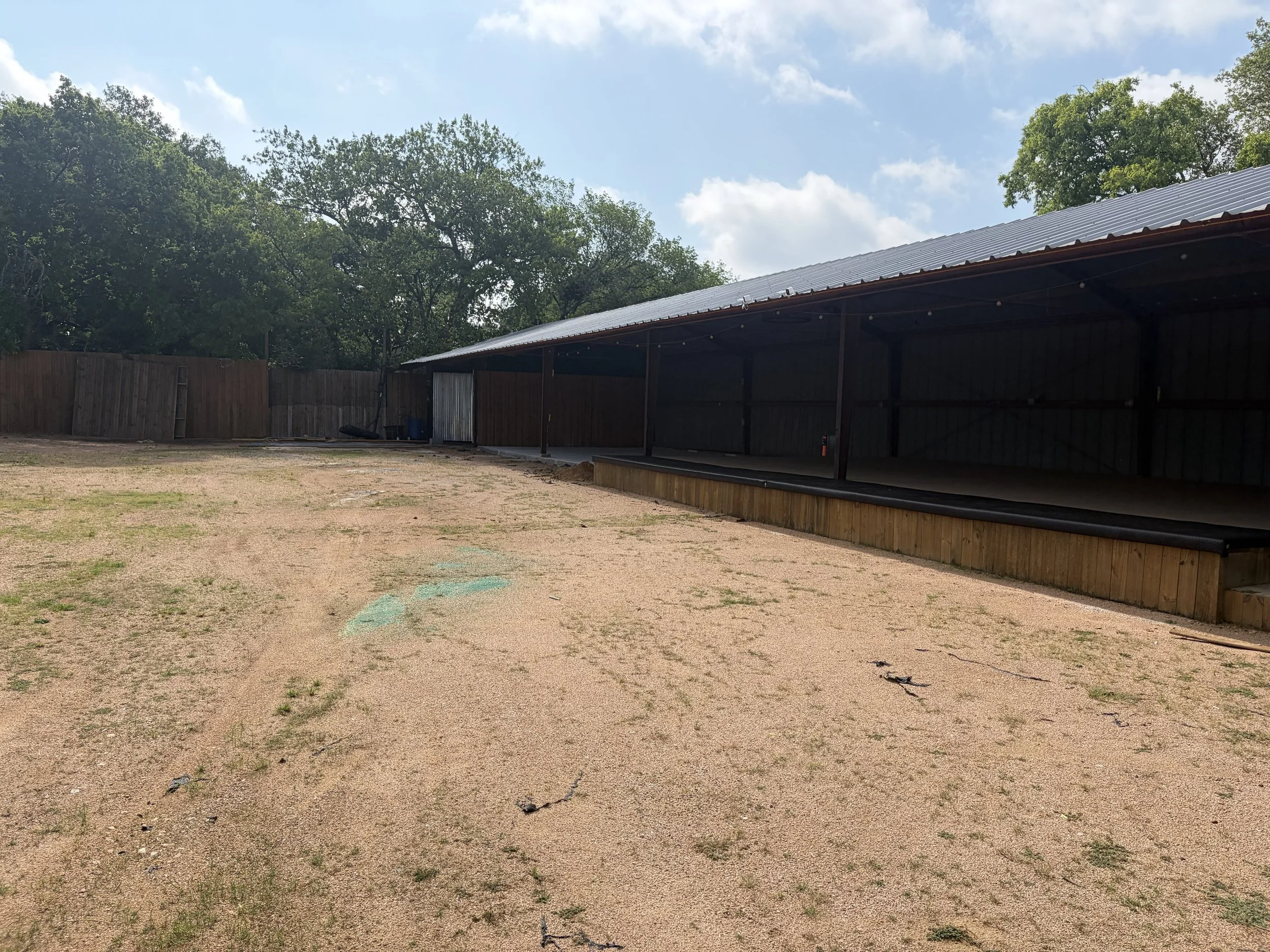 An outdoor scene with a dirt area, a wooden fence, and a metal building with a sloped roof. Trees with green leaves surround the area, and the sky is partly cloudy.