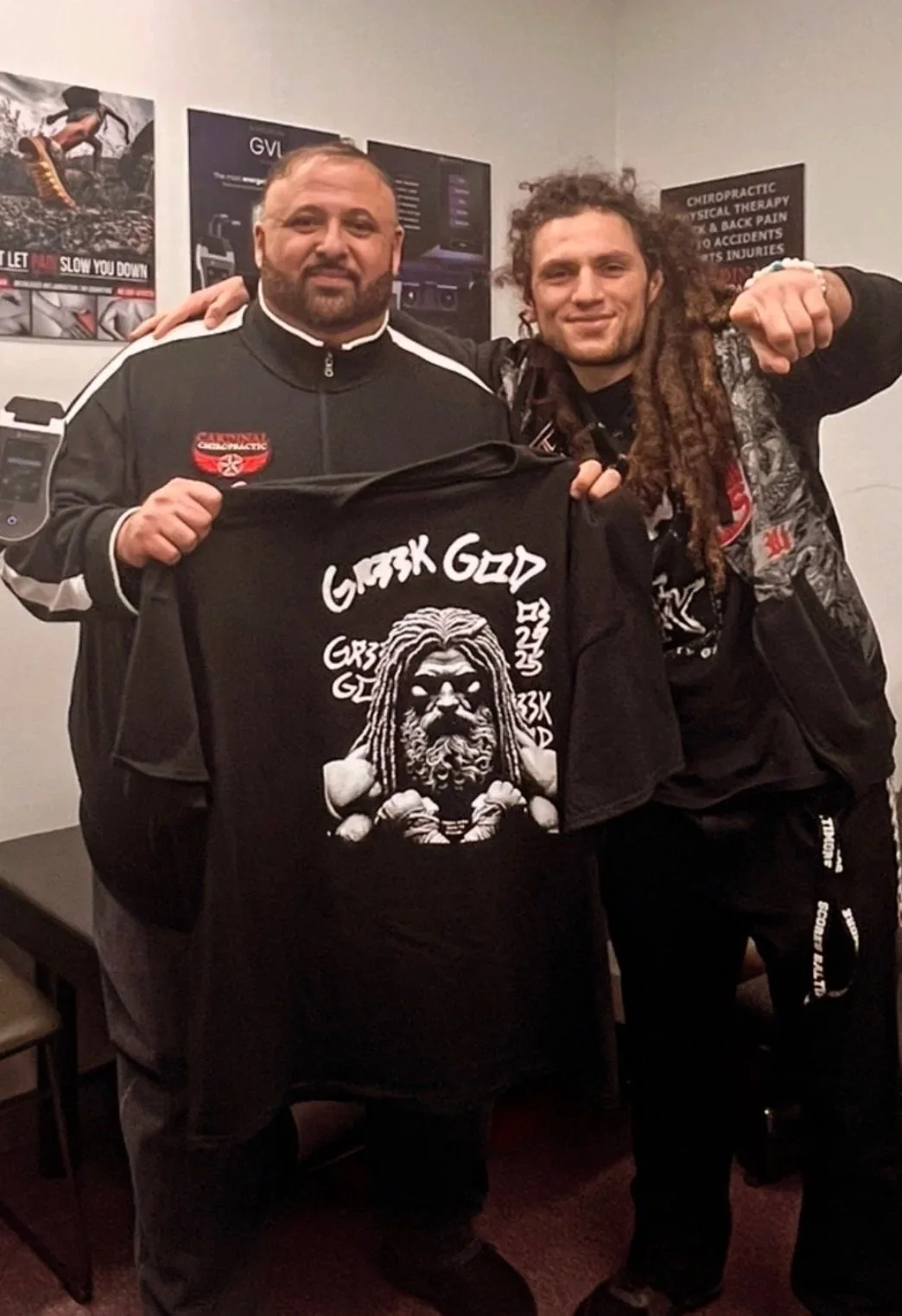Two men posing indoors, one holding a black T-shirt with an illustrated bearded figure with dreadlocks, labeled 'Greek God.' The man on the left has a beard and wearing a black jacket, while the other has long dreadlocks and a patterned jacket. They are smiling and standing close together, with posters and signs visible in the background.