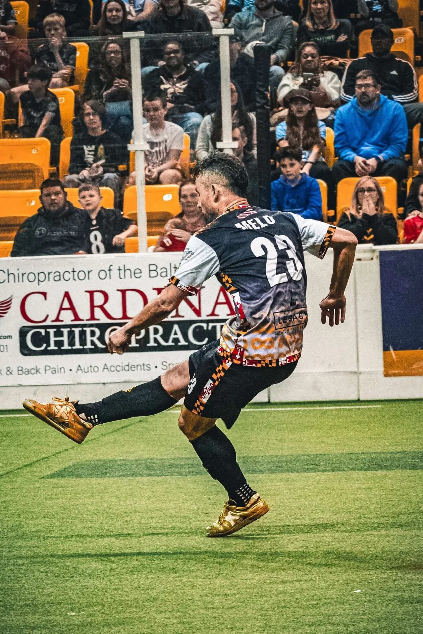 A soccer player wearing a black and white uniform with gold cleats kicks a soccer ball on the field, with a crowd of spectators watching from the stands.