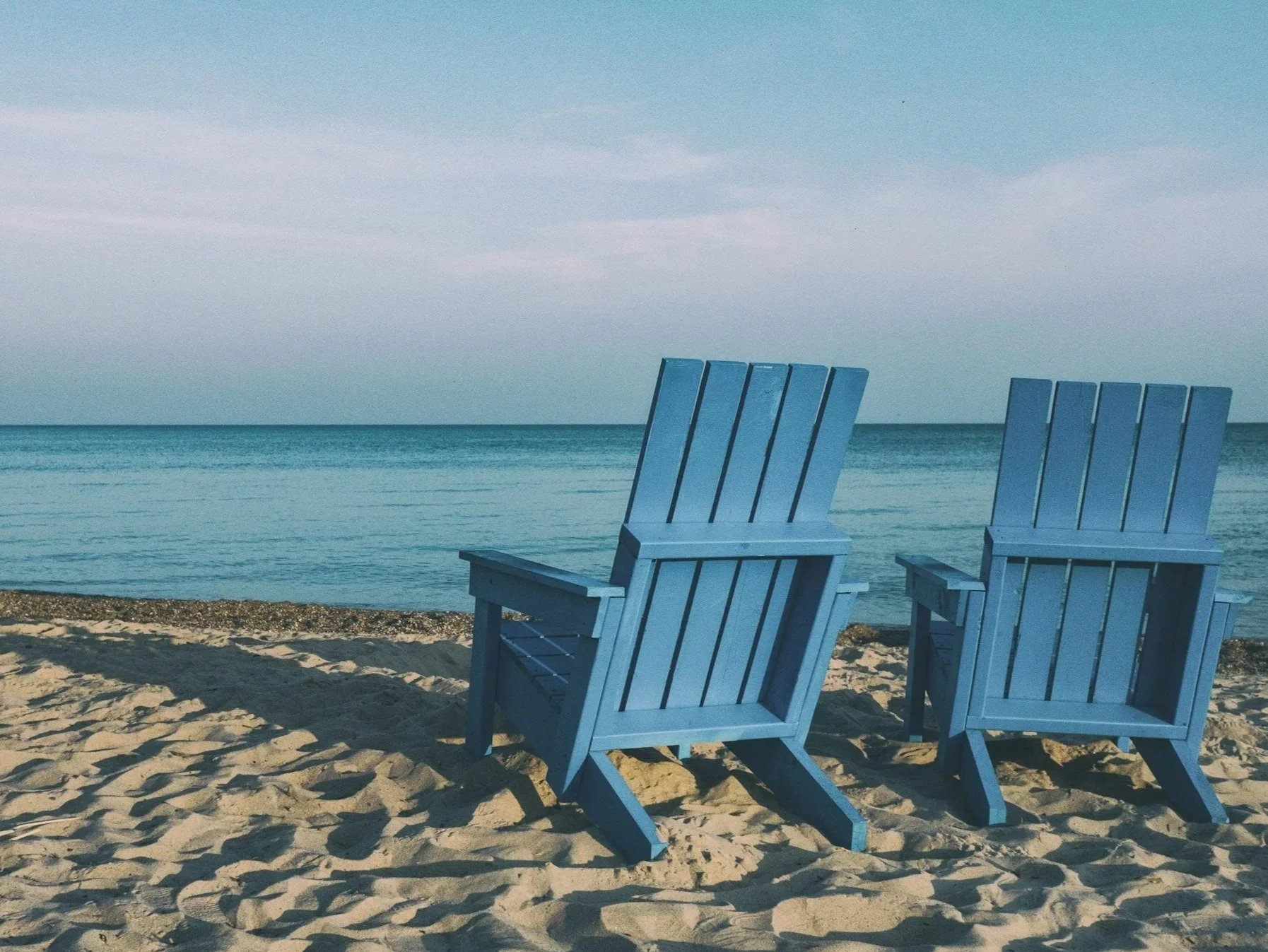 Two blue Adirondack chairs on a sandy beach facing the ocean with calm water and a cloudy sky.