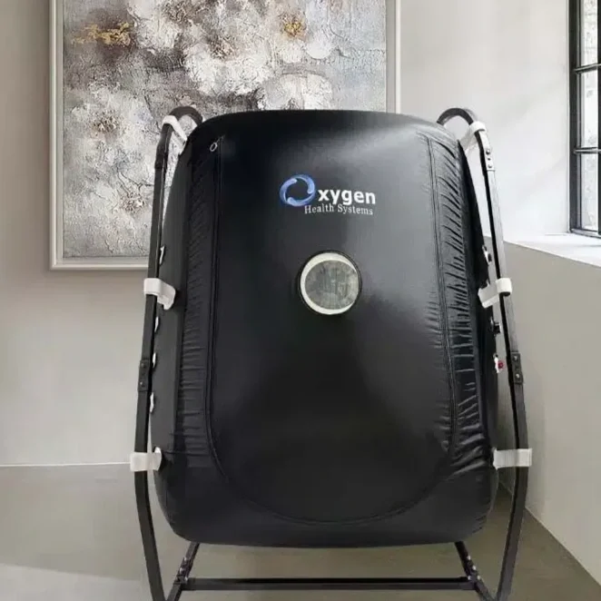 A black hyperbaric oxygen therapy chamber with logo of Oxygen Health Systems, placed indoors against a wall with a window on the right and abstract artwork on the left.