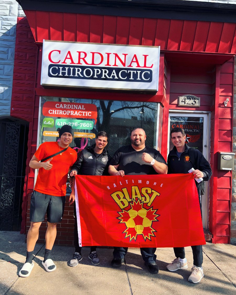 Four men standing in front of a chiropractic office holding a Baltimore Blast banner, with the office sign reading 'Cardinal Chiropractic' and the address 423 visible on the building.