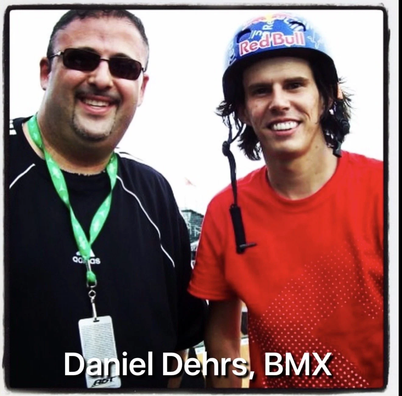 Two men smiling, one wearing sunglasses and sporting a black Adidas shirt with a lanyard, and the other wearing a Red Bull helmet and a red shirt. The text at the bottom reads 'Daniel Dehrs, BMX.'