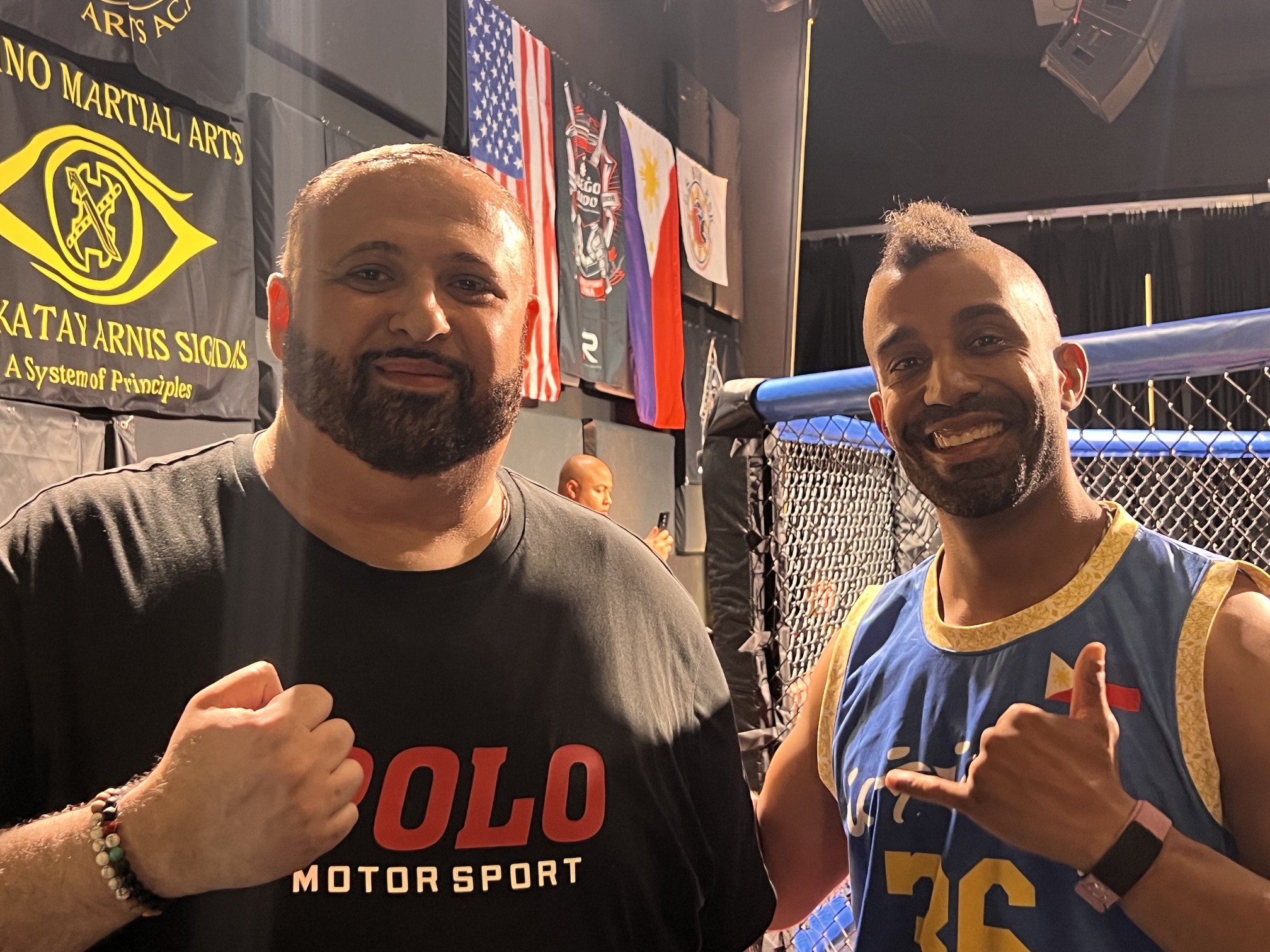 Two men posing inside an MMA or martial arts gym, standing in front of a cage with flags and banners in the background. The man on the left has a beard and wears a black t-shirt with red and white text and wristbands. The man on the right has a mohawk hairstyle, is smiling, and wears a blue and yellow martial arts uniform with a Filipino flag patch. Both are making a fighting stance with clenched fists.