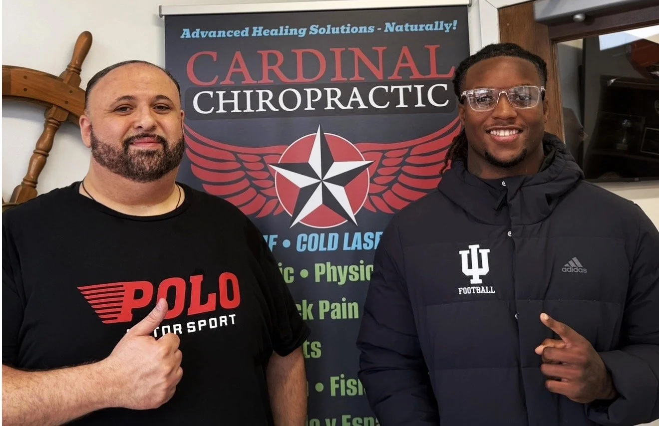 Two men standing in front of a sign for Cardinal Chiropractic. The man on the left is giving a thumbs-up and wearing a black POLO t-shirt. The man on the right is smiling, wearing glasses and a black Indiana University jacket, and pointing at the camera. The sign behind them features a star with red wings and lists services like advanced healing solutions, cold laser, chiropractic, physical therapy, back pain, sports, fish oil, and more.