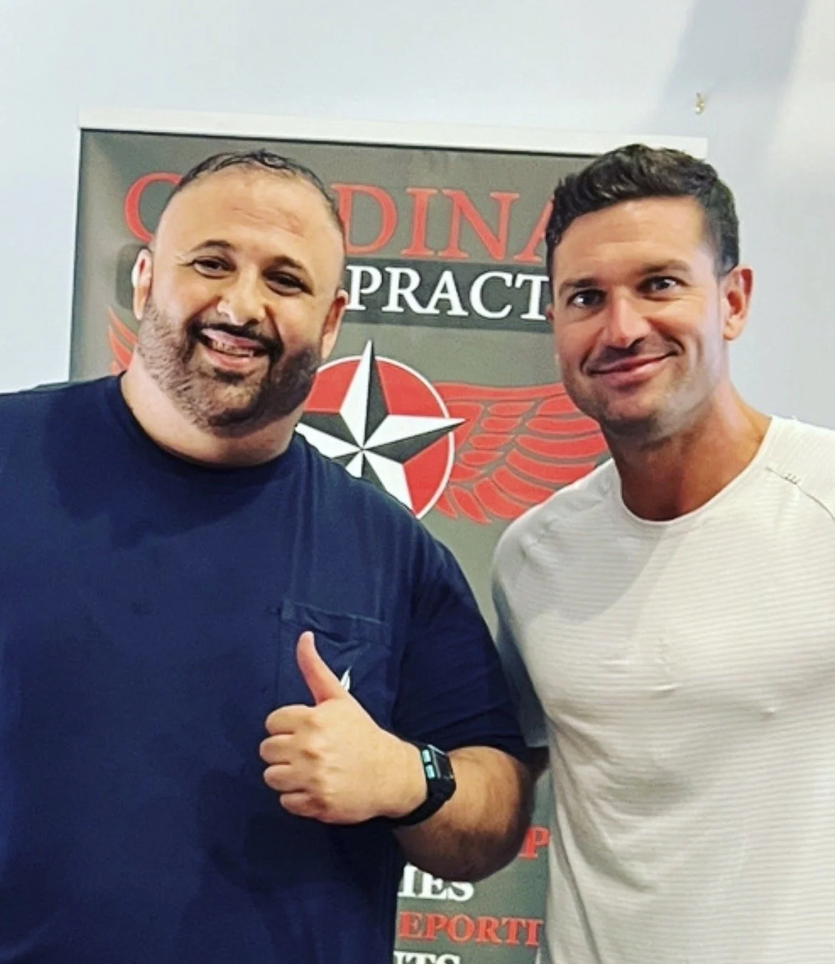 Two men standing together indoors, smiling at the camera, with a sign in the background that reads 'Chiropractic Practice' and has a star and wings logo.