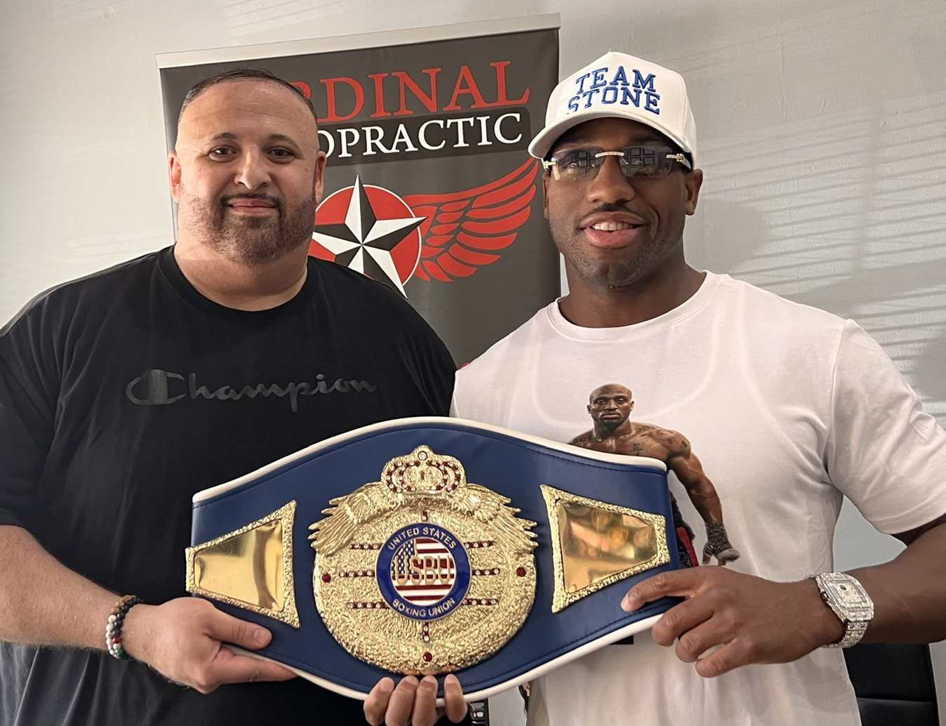 Two men holding a United States Boxing Union championship belt, standing in front of a sign that says 'CARDINAL CHIROPRACTIC'. One man is wearing a black champion t-shirt, and the other is wearing a white t-shirt with a boxer image. The man on the right is also wearing glasses, a white cap, a watch, and a bracelet.