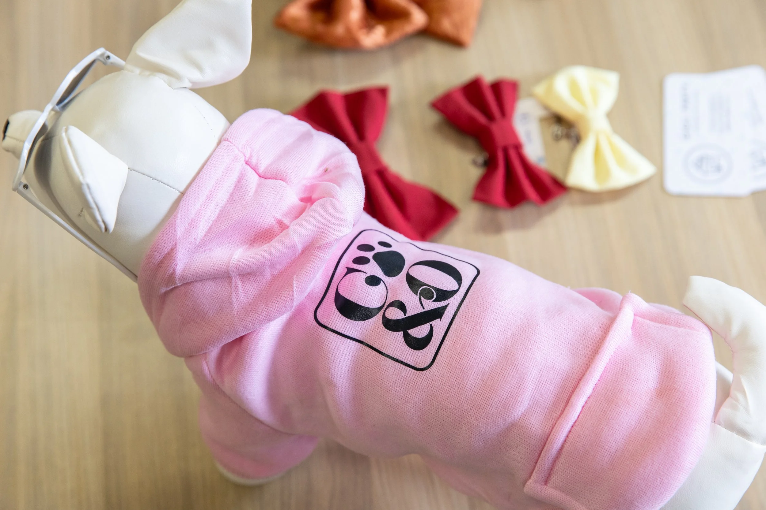 A stuffed dog wearing a pink hoodie with a paw print and the word 'Joe' on it, surrounded by colorful fabric bows on a wooden surface.