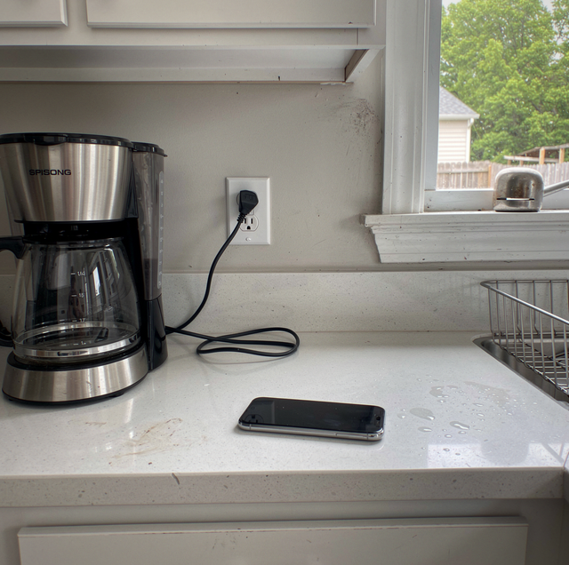 A coffee maker sits on a kitchen counter next to a smartphone and power cable by a window.
