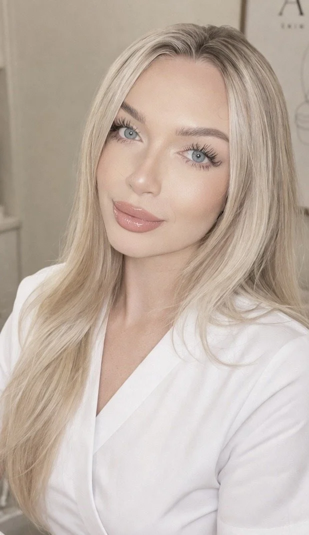 Close-up of a woman with long blonde hair, blue eyes, and glossy lips, wearing a white top, in an indoor setting.