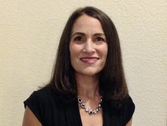 A woman with shoulder-length dark brown hair, smiling, wearing a black top and a silver necklace with purple stones, standing against a beige wall.