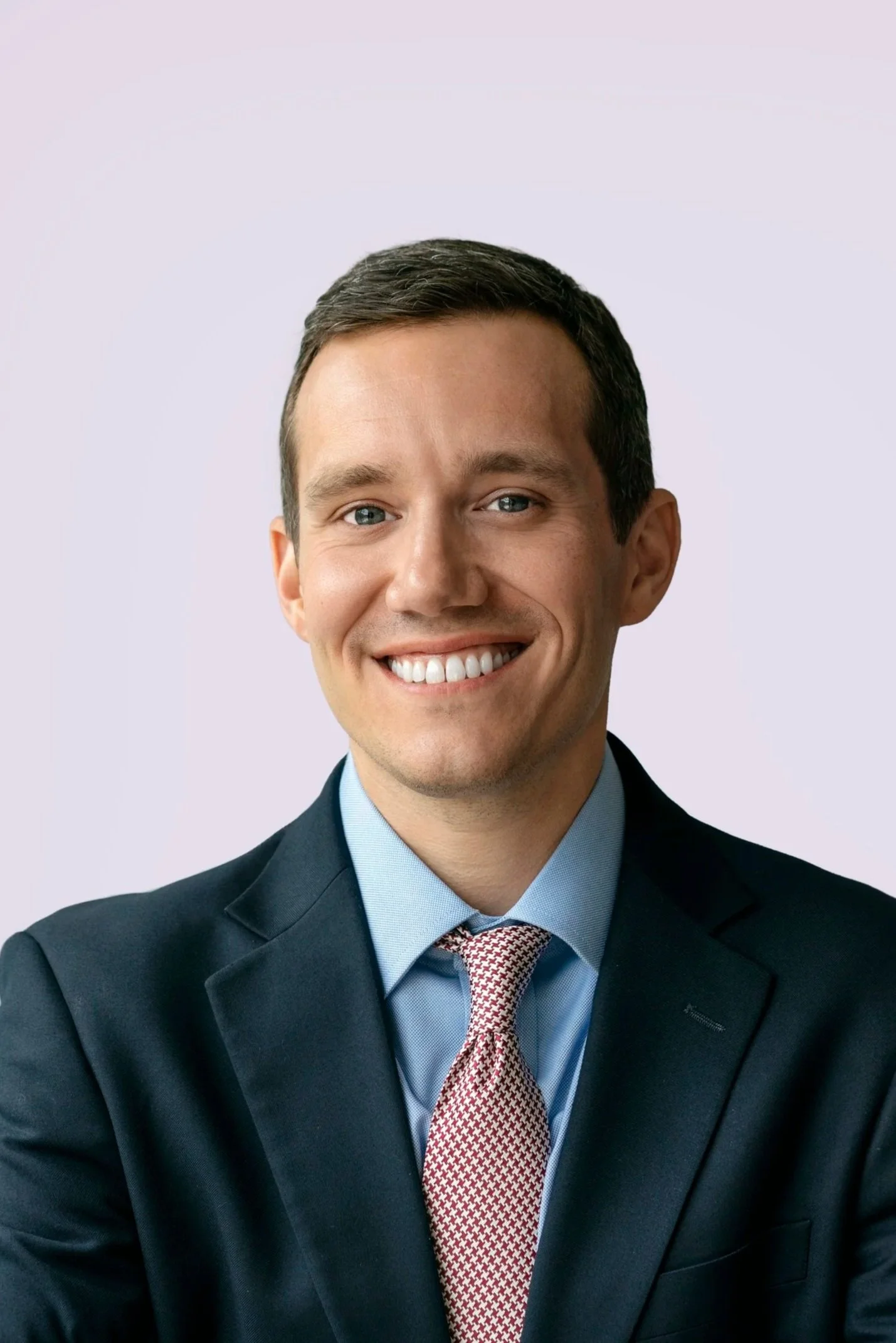 A smiling man wearing a dark business suit, light blue shirt, and red patterned tie, posing against a plain light background.