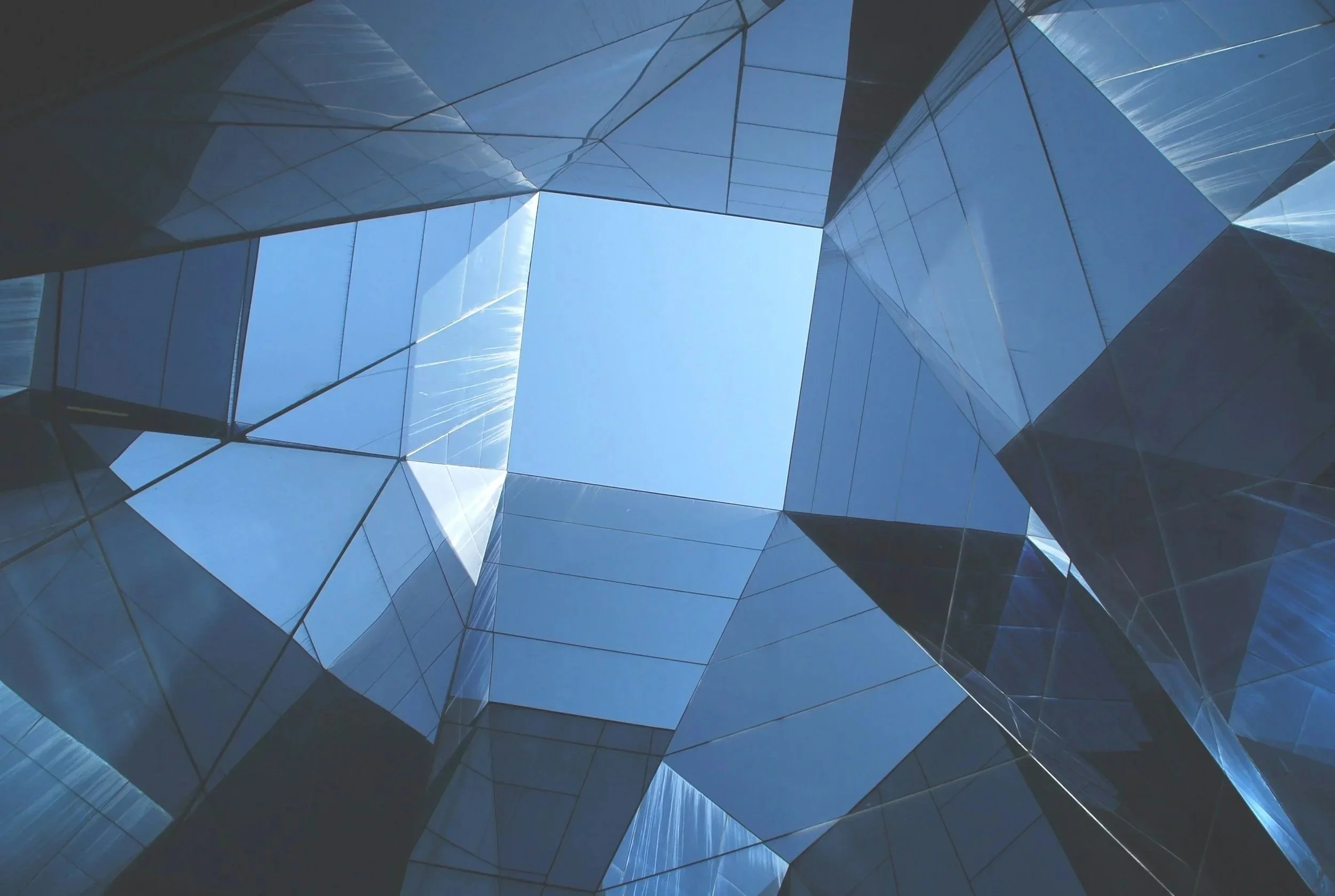 Looking up at a modern glass building with reflective surfaces and a clear blue sky visible through an opening at the top.