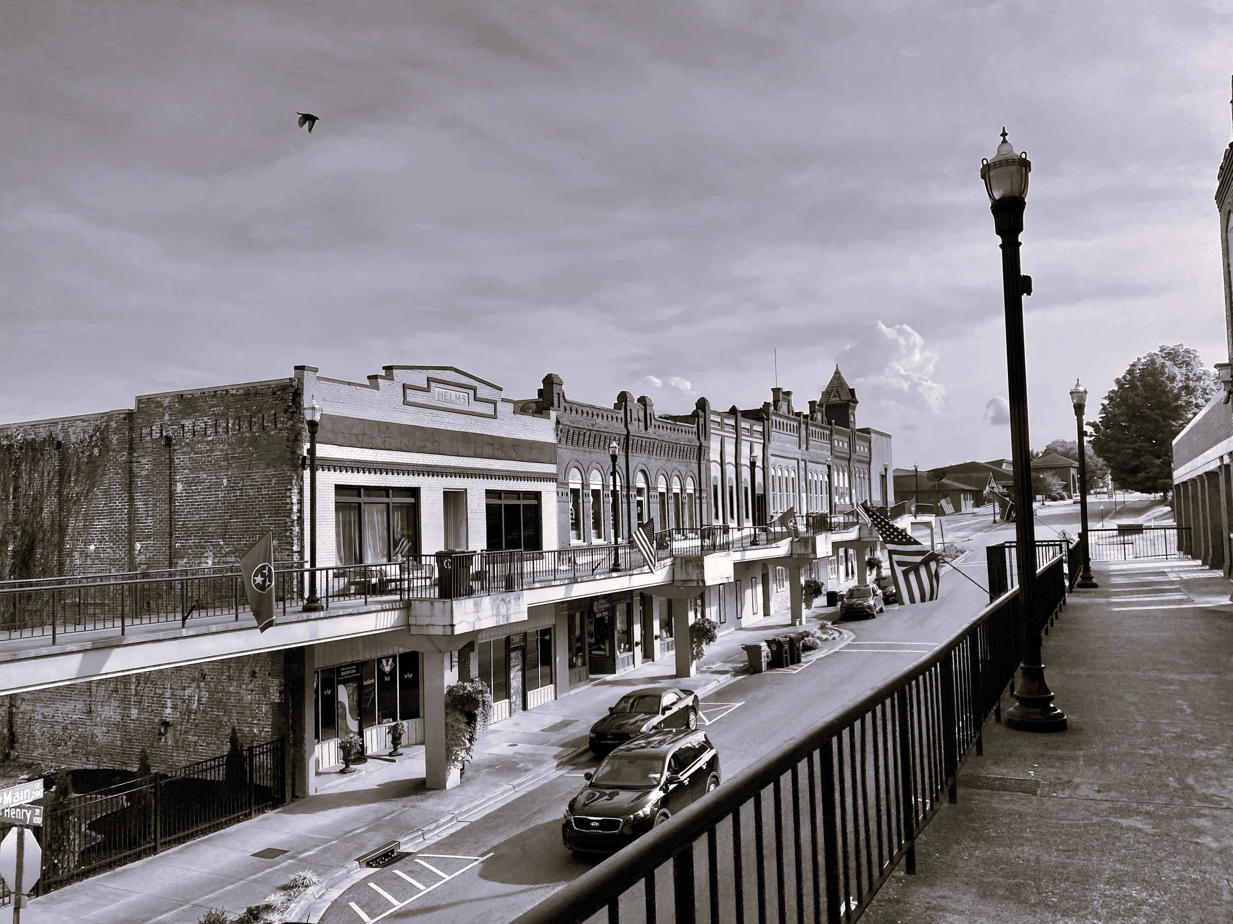 Black and white photo of a small town street with historic buildings, American flags, a sidewalk with lampposts, and parked cars.