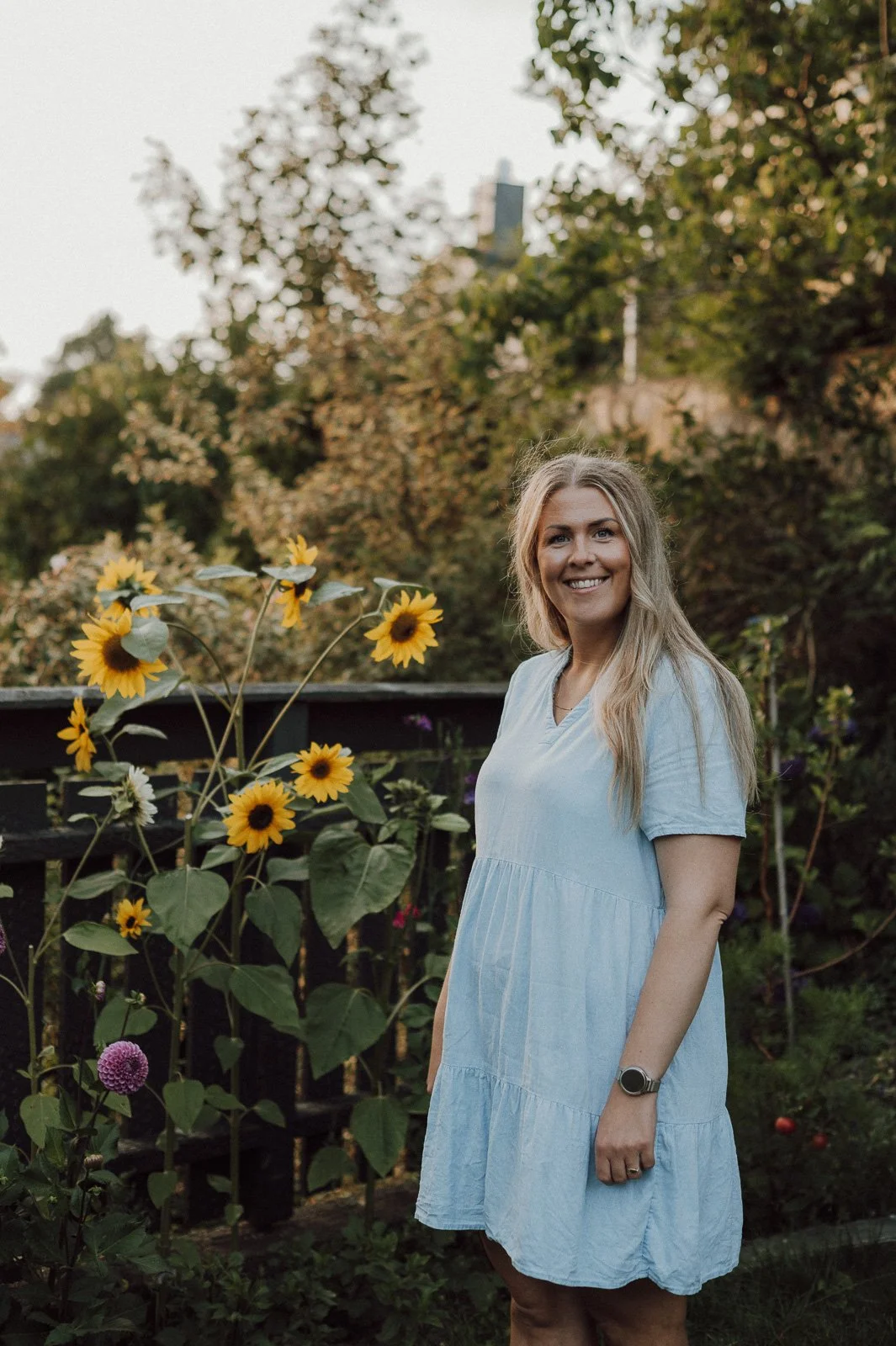 En kvinne i en sommerkjole står i en hage med blomster, med trær i bakgrunnen, og smiler mot kameralinsen.