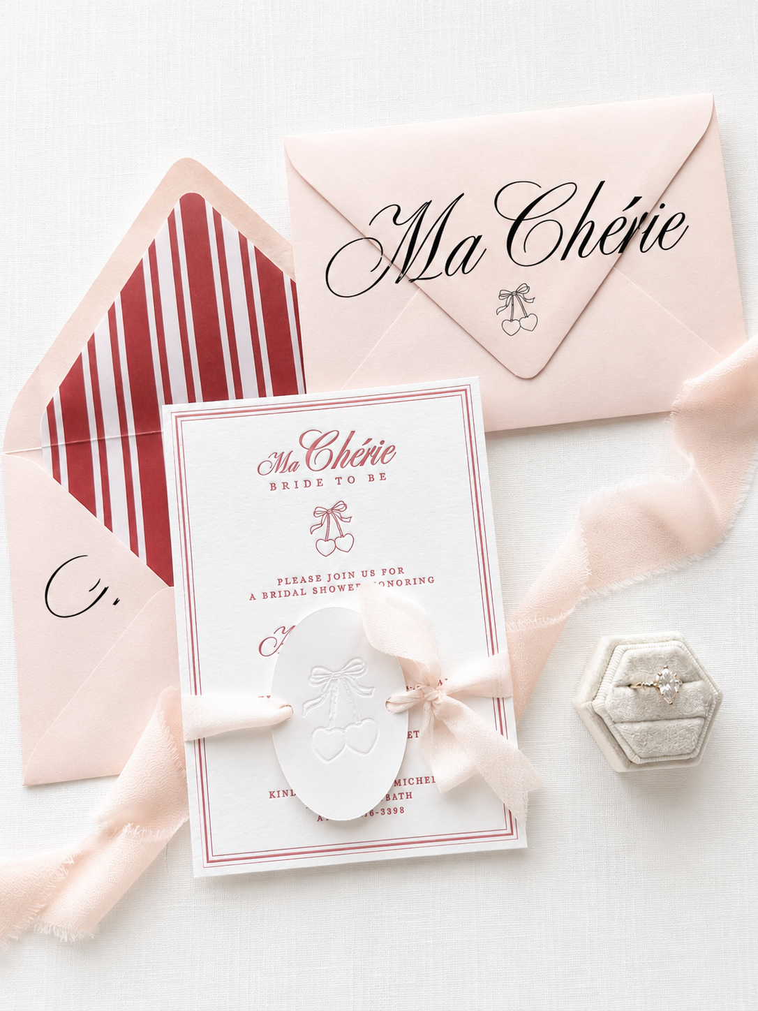 Invitation to a bridal shower with pink and white theme, featuring a card with cherries and ribbon graphic, a pink envelope labeled "Ma Chérie," a ring in a velvet box, and pink ribbon.
