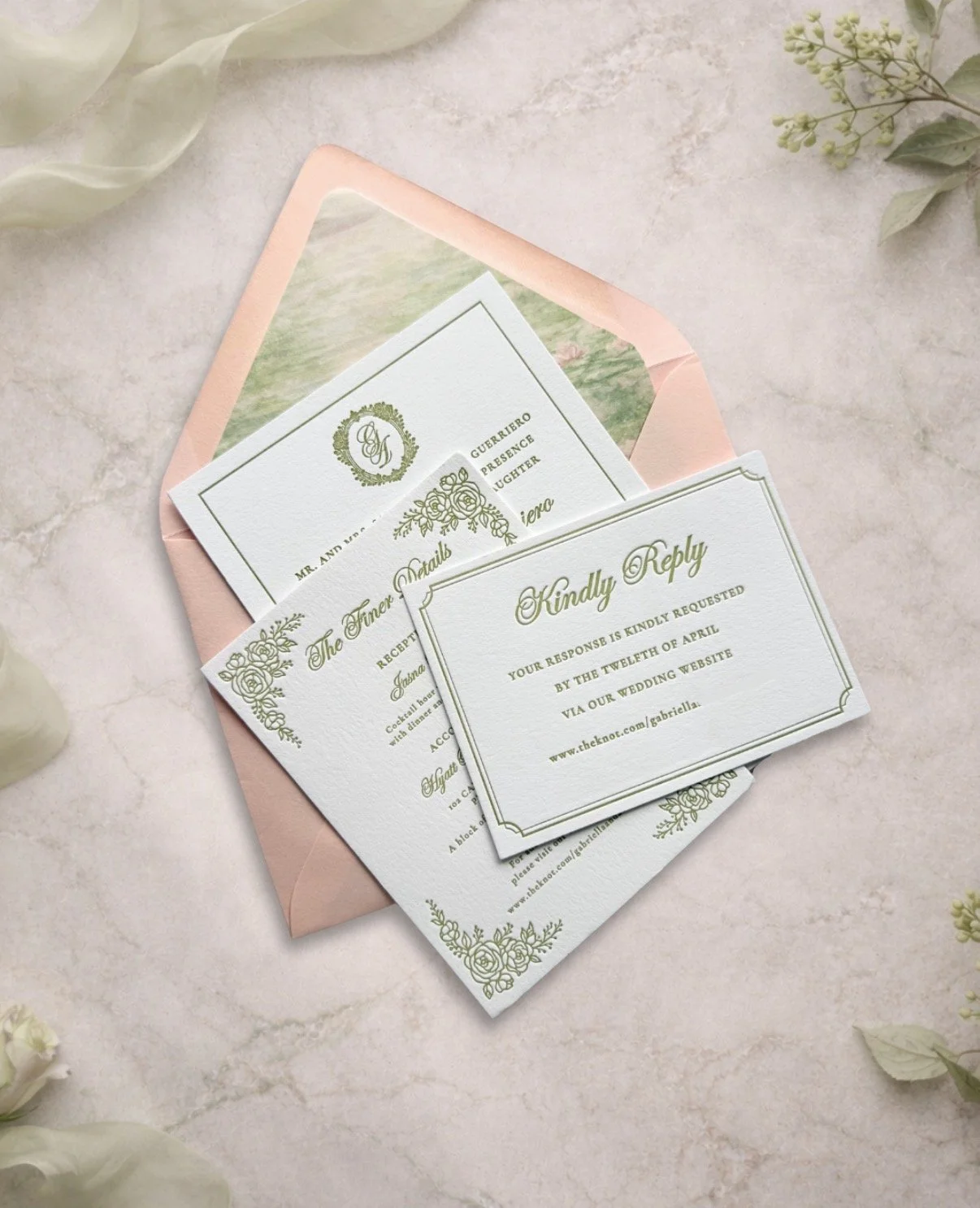 Elegant wedding RSVP and details cards with gold accents, placed on a pink envelope and surrounded by white flowers and greenery.