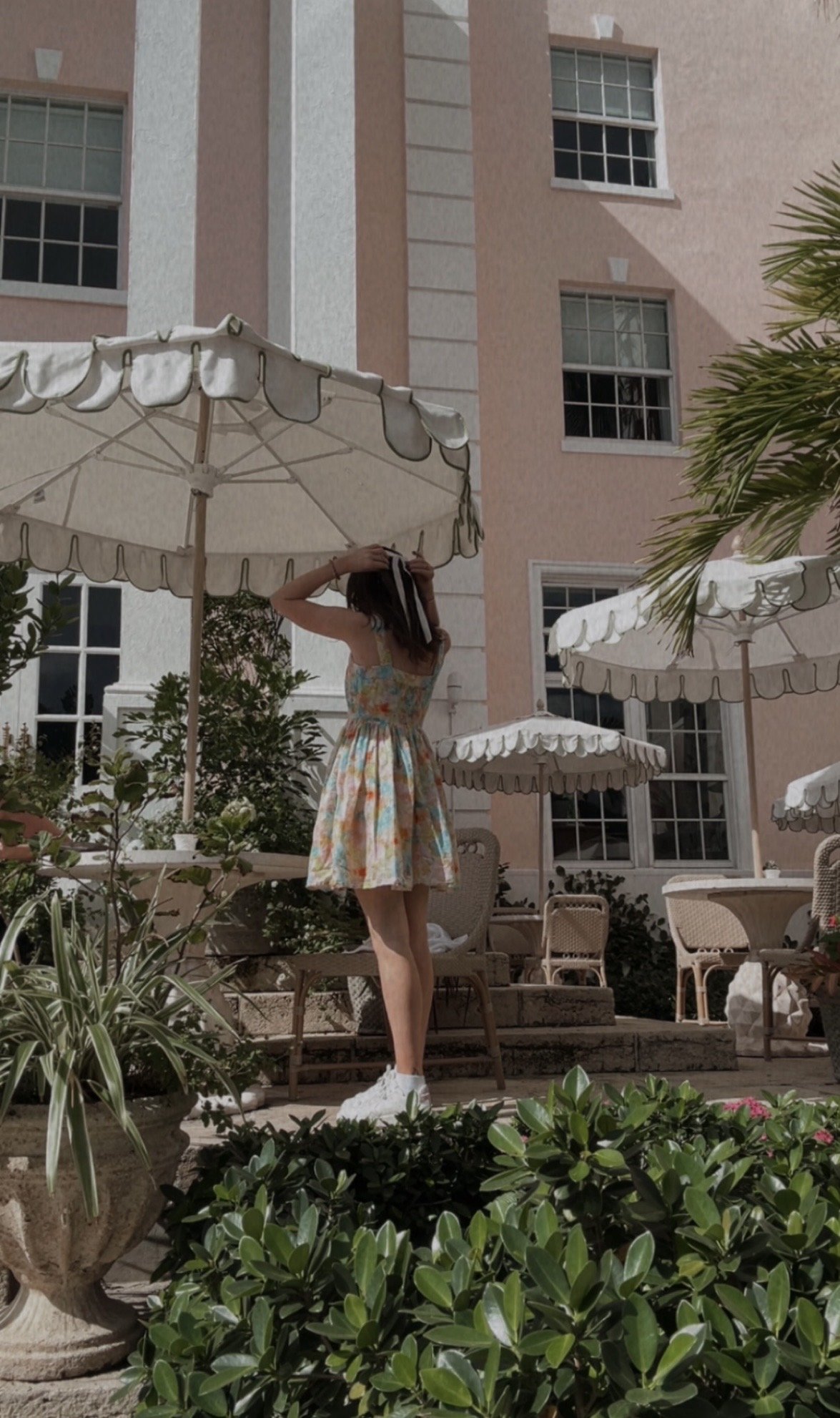 A woman in a colorful dress standing outdoors in front of a pink building with large windows, surrounded by patio tables, chairs, umbrellas, and green plants.