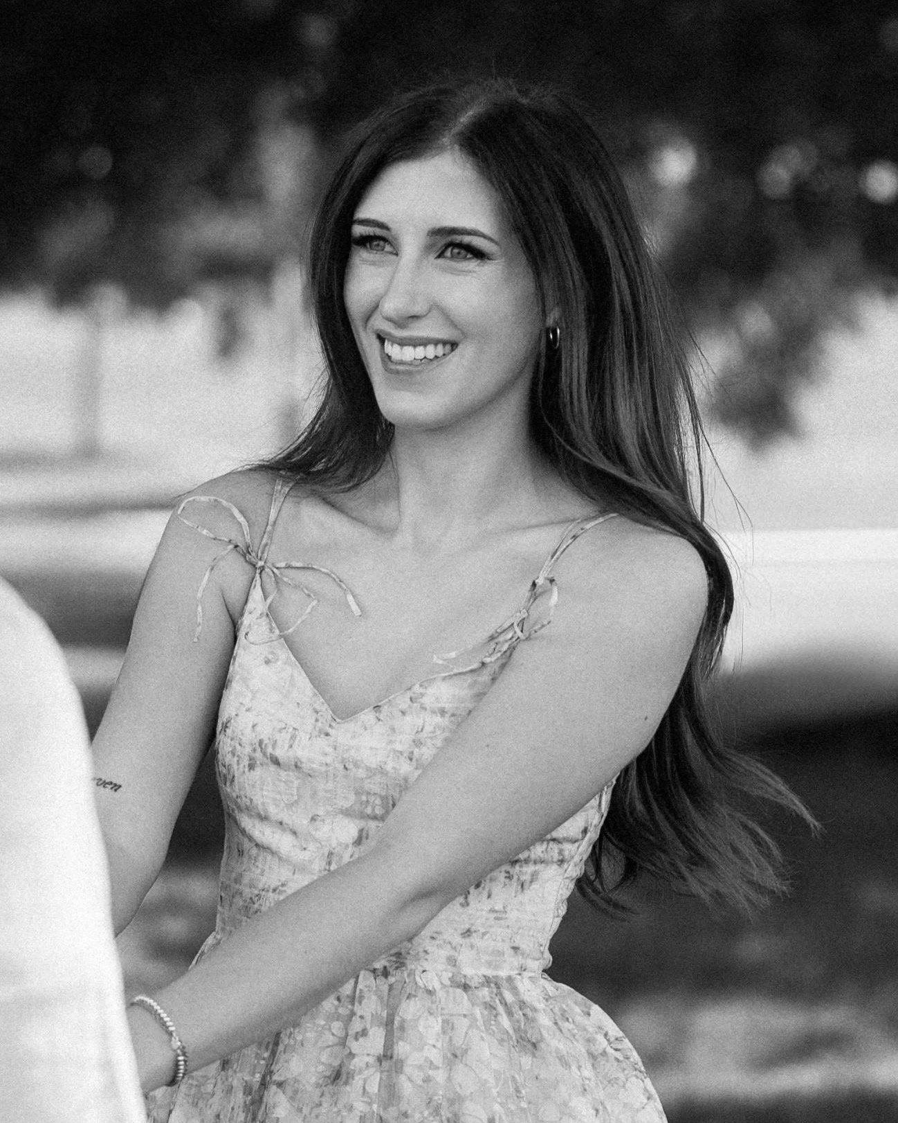 A woman with long dark hair smiling outdoors, wearing a sleeveless dress with tied straps on the shoulders.