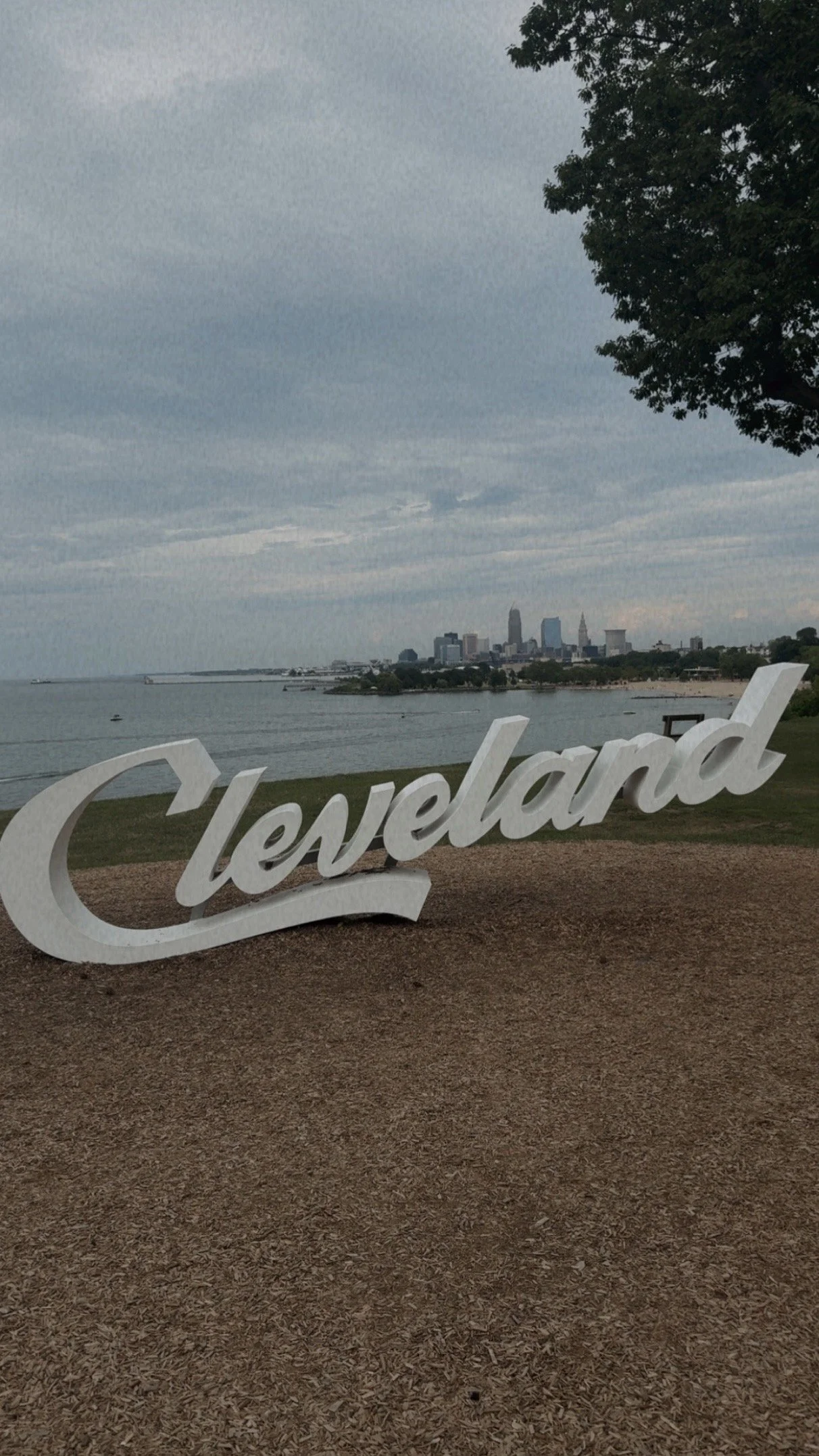 A large white decorative sign spelling 'Cleveland' is placed on a dirt path near a body of water, with the city skyline visible in the distance against a cloudy sky.