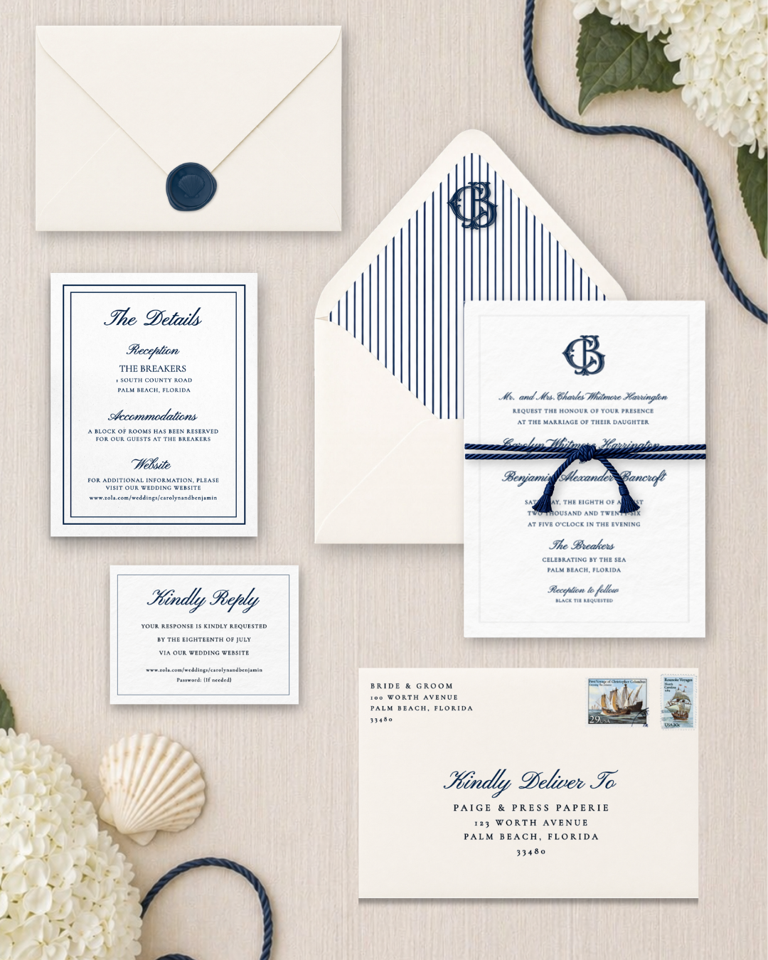Wedding invitation suite, including an envelope with a blue wax seal, a reception and accommodation card, an invitation card tied with blue cord, and mailing details, all arranged on a white wooden surface with flowers and seashells.